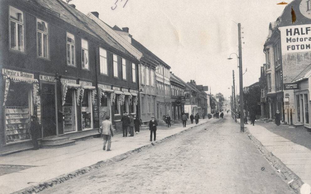 Et vintage gatebilde med folk som går og butikker på begge sider, inkludert en bokhandel og en butikk med motortilbehør. Gaten er ikke asfaltert og kantet med bygninger i urbane omgivelser.