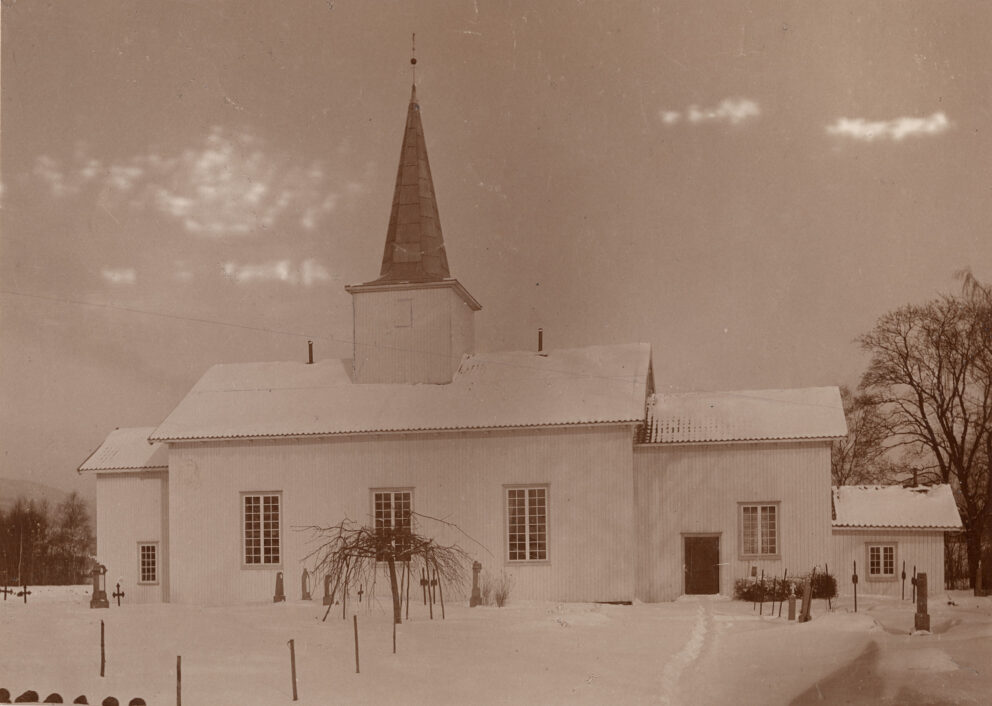 Sepia-tonet bilde av en kirke med et høyt tårn, omgitt av snø. Det er nakne trær og noen få graver synlige i forgrunnen.