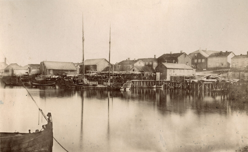 Et sepia-tonet bilde viser en historisk havn med flere seilbåter som ligger til kai i nærheten av trebygninger og en brygge, og reflekterer over stille vann.