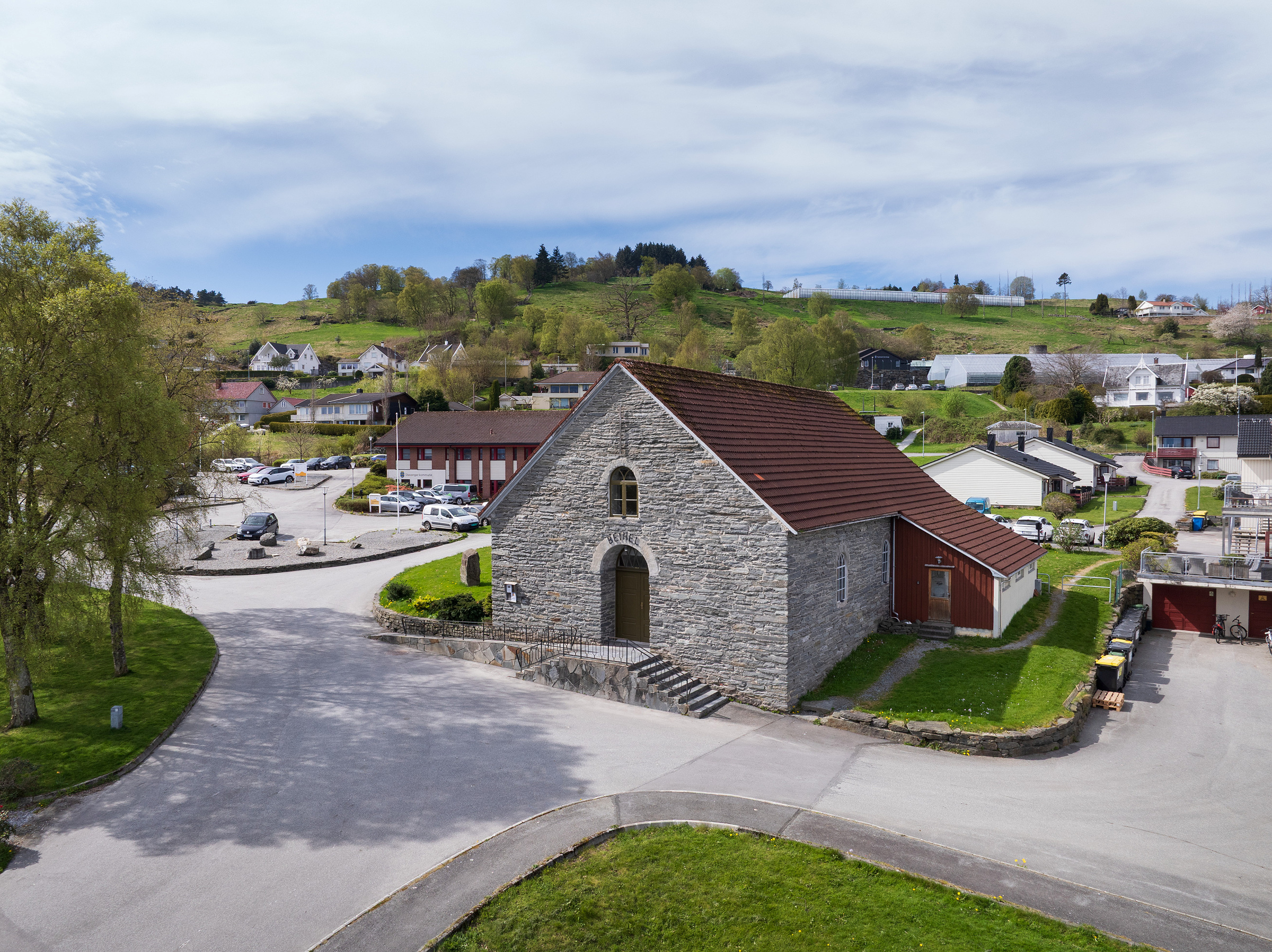 En steinkirke med brunt tak står ved et gatekryss i en liten by med spredte hus og grøntområder i en åsside i bakgrunnen.