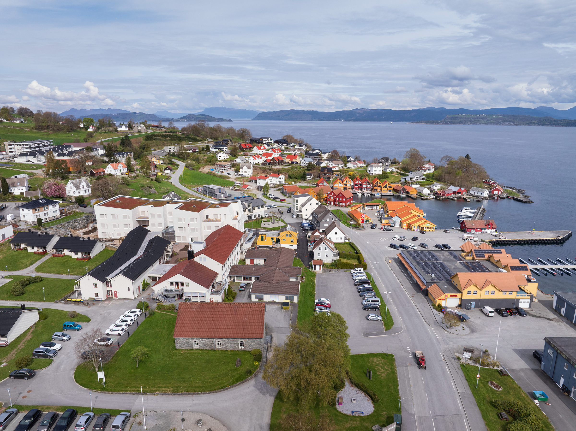 Luftfoto av en kystlandsby med hus, parkerte biler, en havn og omkringliggende grøntområder ved siden av et vann under en delvis overskyet himmel.