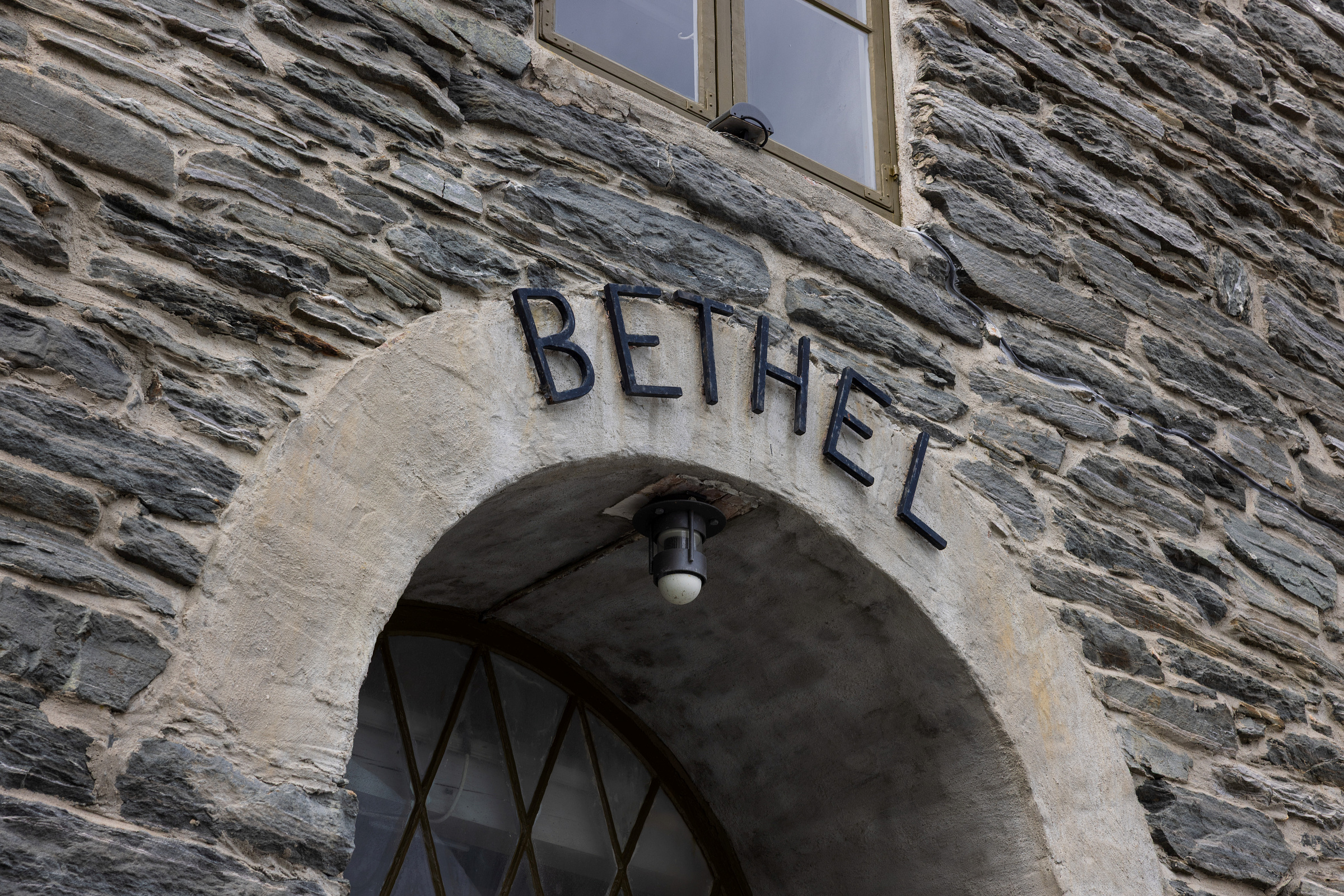 Fasade av steinbygning med en buet inngang, over hvilken ordet BETHEL er vist med svarte metallbokstaver. En lysarmatur henger fra buen.