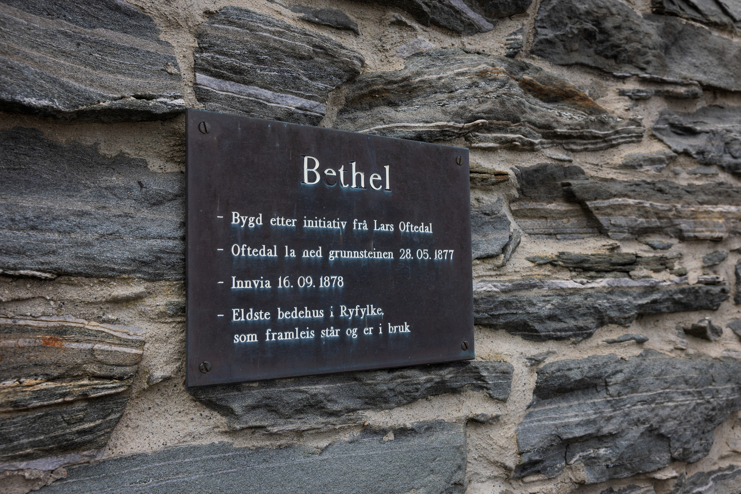En metallplakett på en steinmur viser norsk tekst om Betel, som beskriver grunnleggelsesdatoene og den historiske betydningen i Ryfylke.