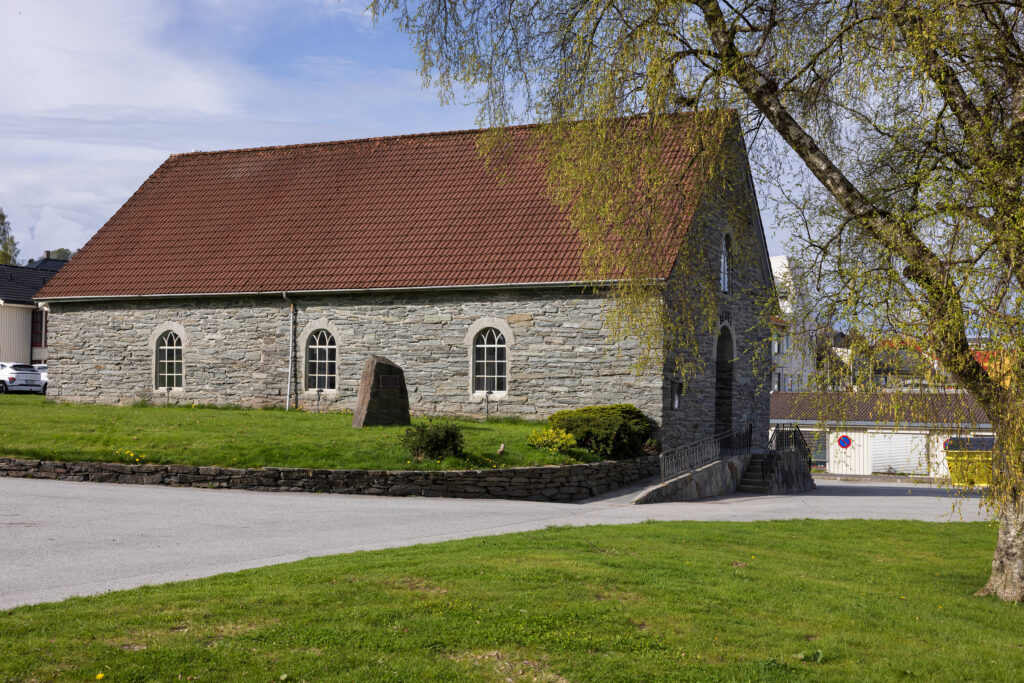 En steinkirke med buede vinduer og et rødt tegltak står på en gresskledd tomt med et tre og et lite steinmonument foran.