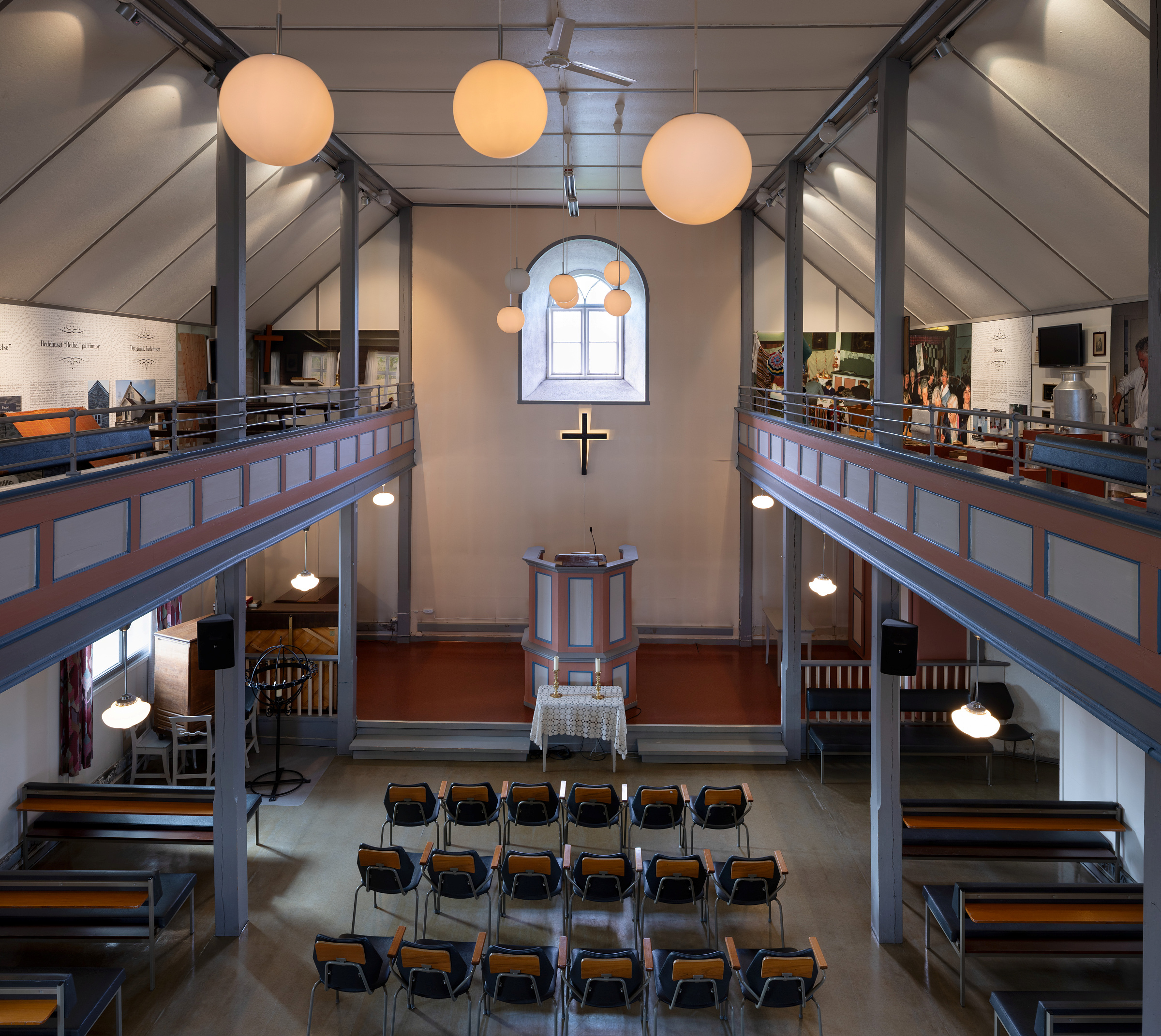 Interiør i en kirke med rader med tomme stoler vendt mot en prekestol, et kors på veggen og to sidebalkonger, opplyst av runde taklamper.