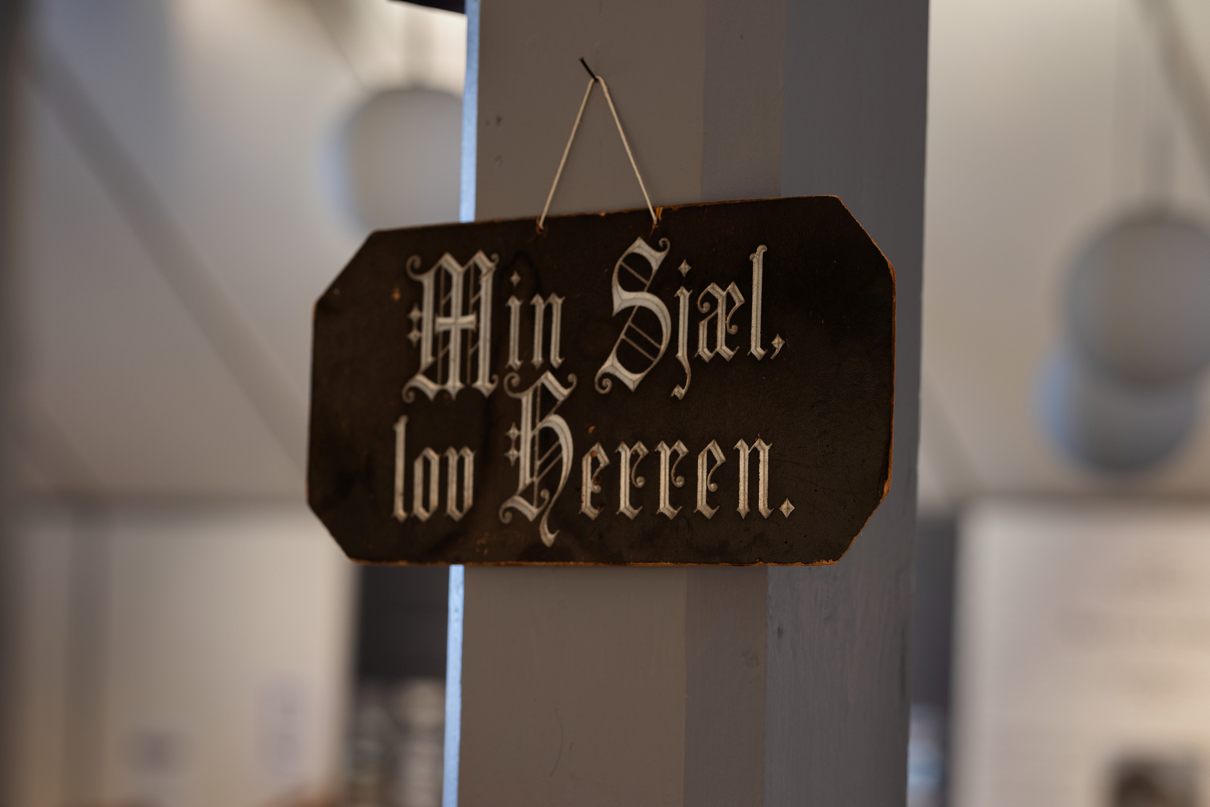 Et svart skilt med hvit gotisk skrift, hengende på en spiker, lyder «Min Sjal, lov Herren.» innendørs.