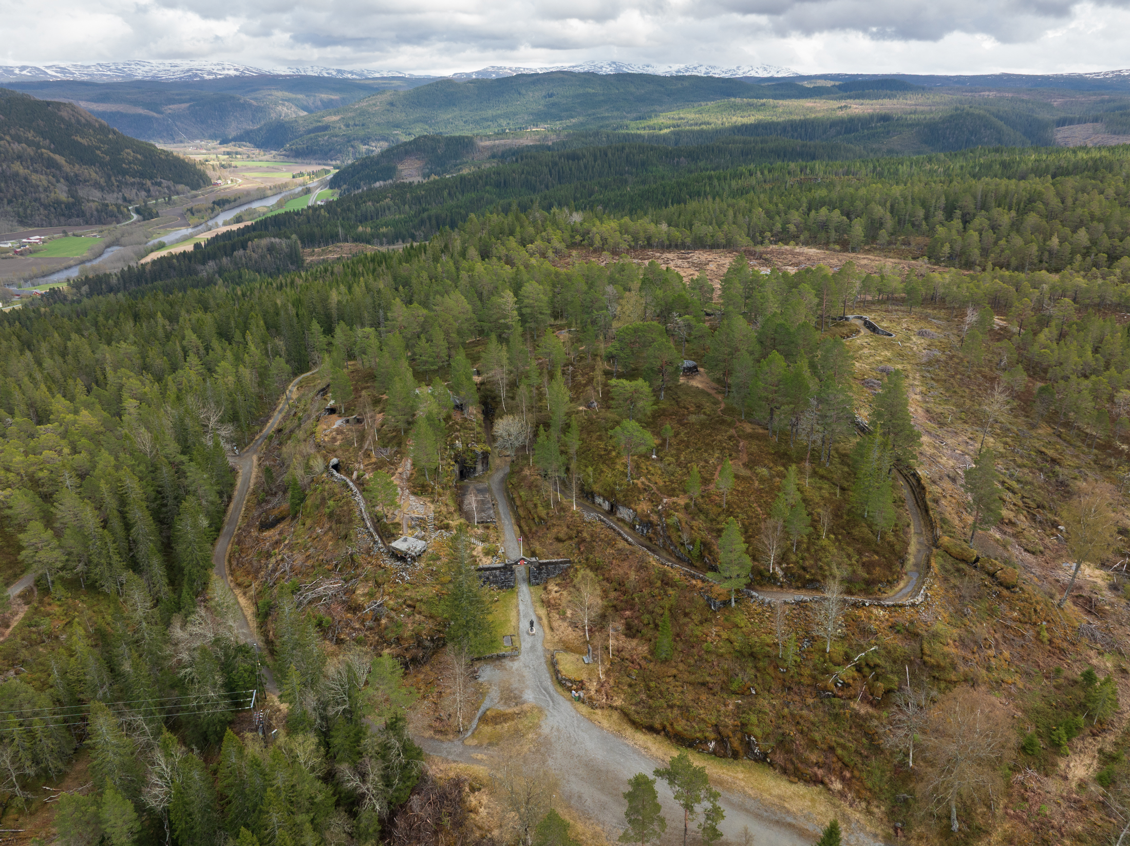 Luftfoto av et kupert, skogkledd landskap med spredte ruiner og steinmurer, gjennomskåret av grusstier og omgitt av fjell under overskyet himmel.