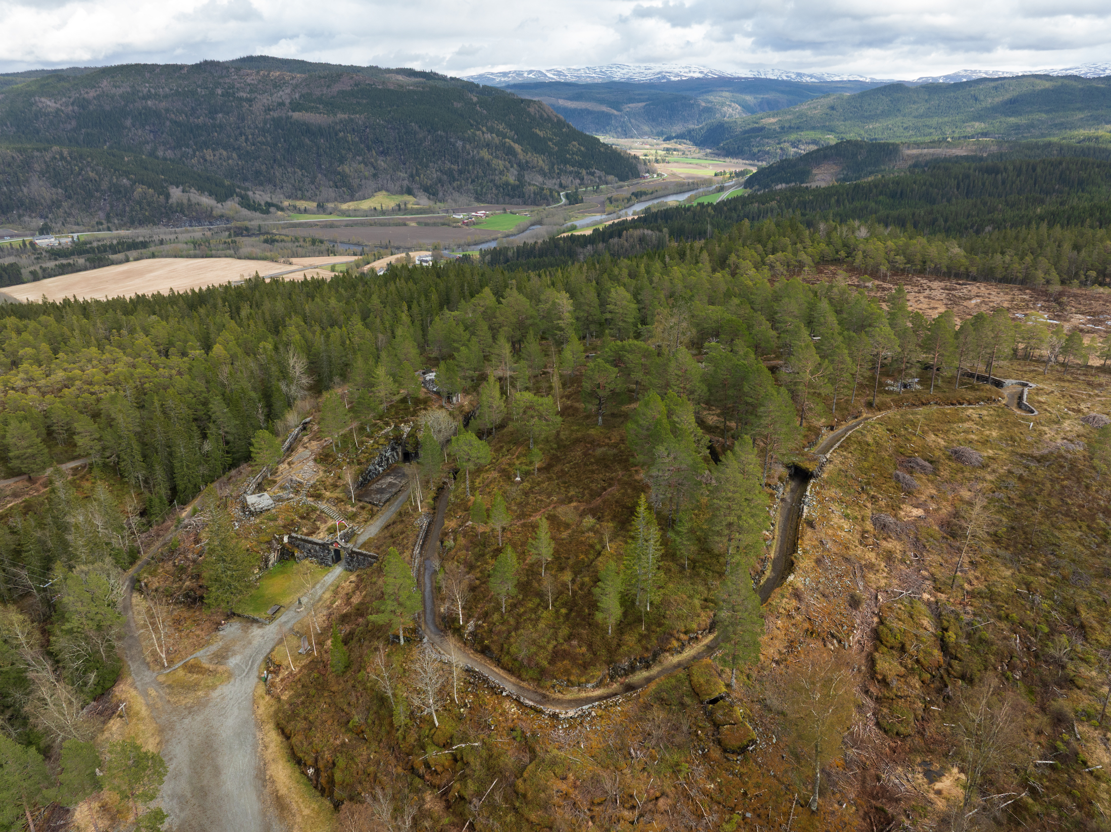Luftfoto av et Hegra festning med turstier og noen små steinstrukturer, omgitt av åser og fjerntliggende jordbruksland under en overskyet himmel.