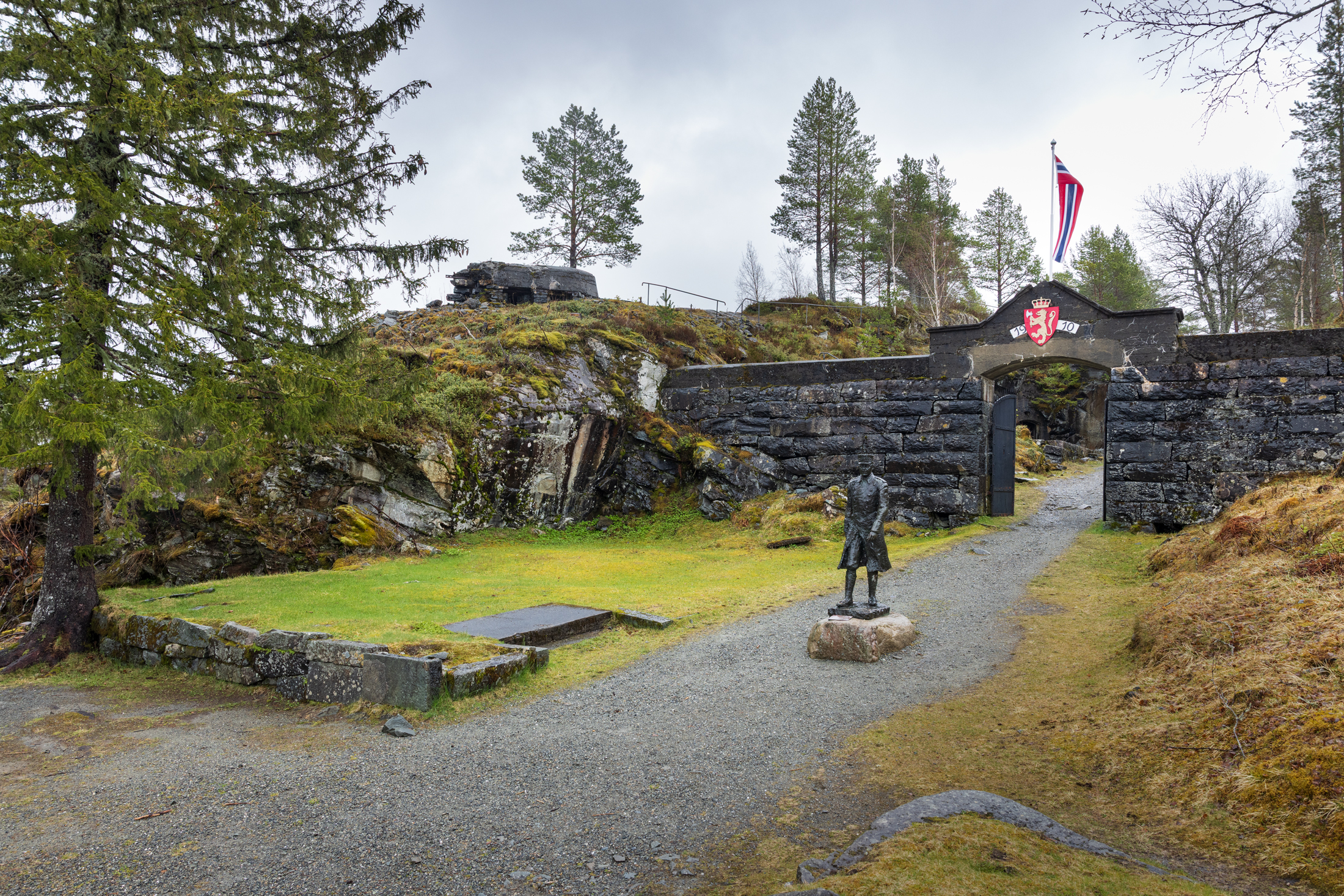 En statue av major Hans Reidar Holtermann står foran en Hegra festning med våpenskjold og norsk flagg, omgitt av trær og gresskledd terreng.
