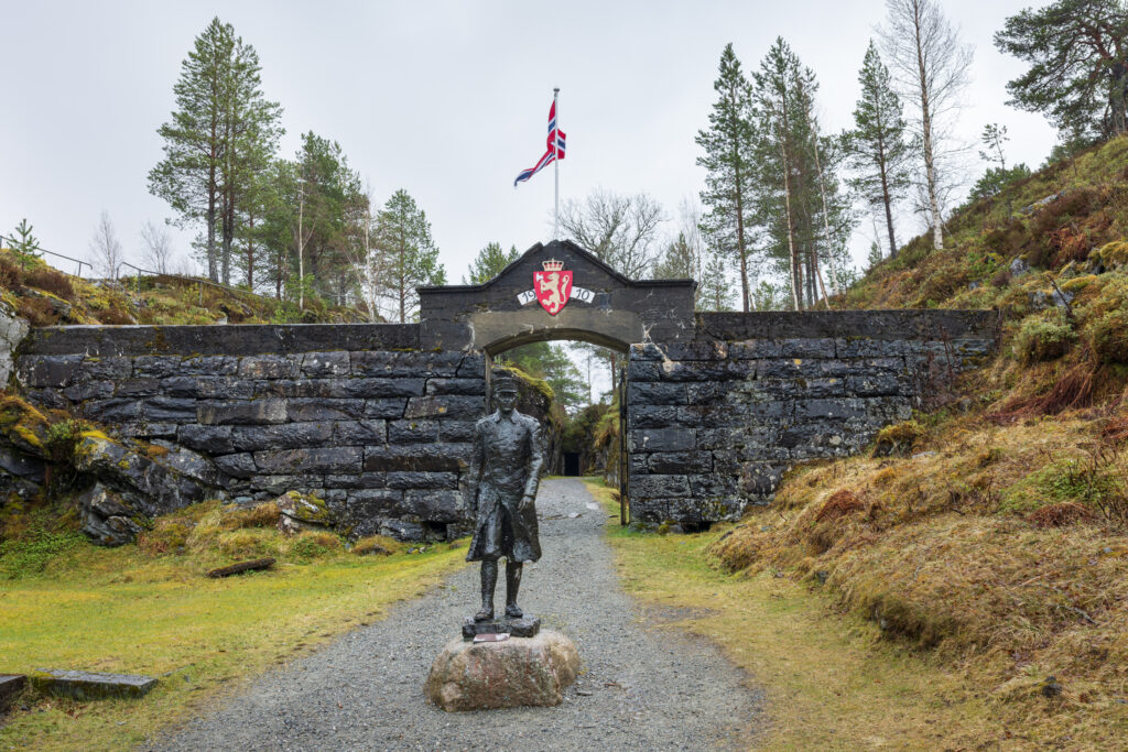 En statue av major Hans Reidar Holtermann står foran en Hegra festning med våpenskjold og norsk flagg, omgitt av trær og gresskledd terreng.