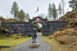 En statue av major Hans Reidar Holtermann står foran en Hegra festning med våpenskjold og norsk flagg, omgitt av trær og gresskledd terreng.