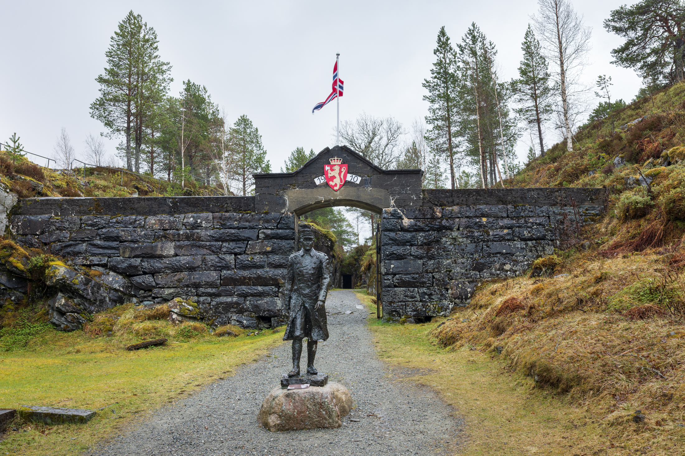 En statue av major Hans Reidar Holtermann står foran en Hegra festning med våpenskjold og norsk flagg, omgitt av trær og gresskledd terreng.