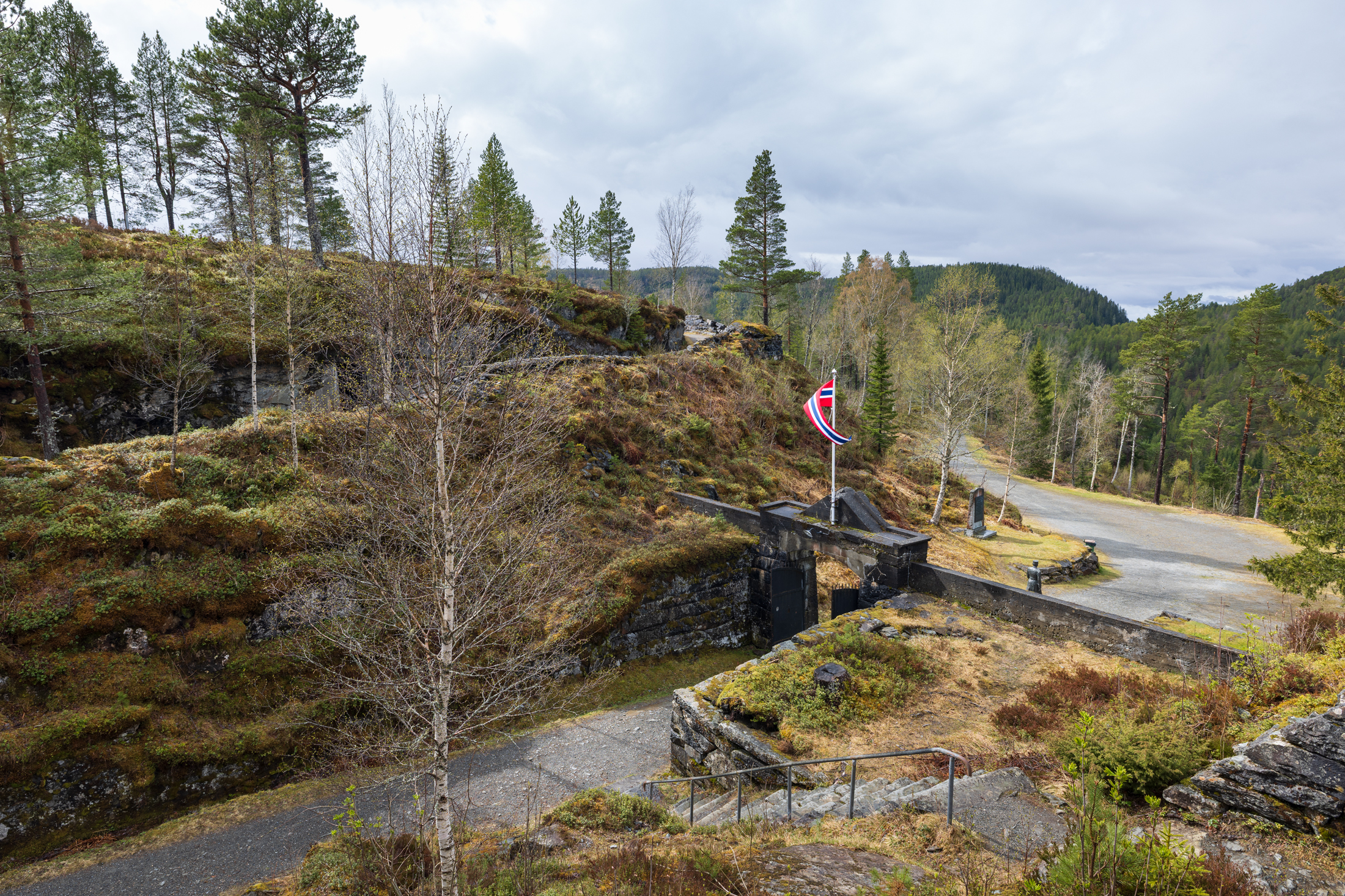 Hegra festning med et norsk flagg står blant trær og steinete terreng, med en asfaltert sti og skogkledde åser i bakgrunnen.