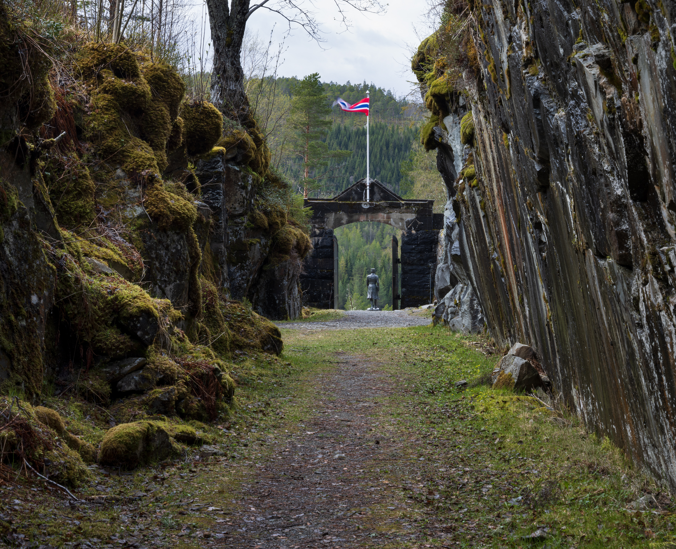 En grussti mellom mosegrodde fjellvegger fører til en treport med et norsk flagg på toppen; en statue står utenfor porten i et skogkledd område.