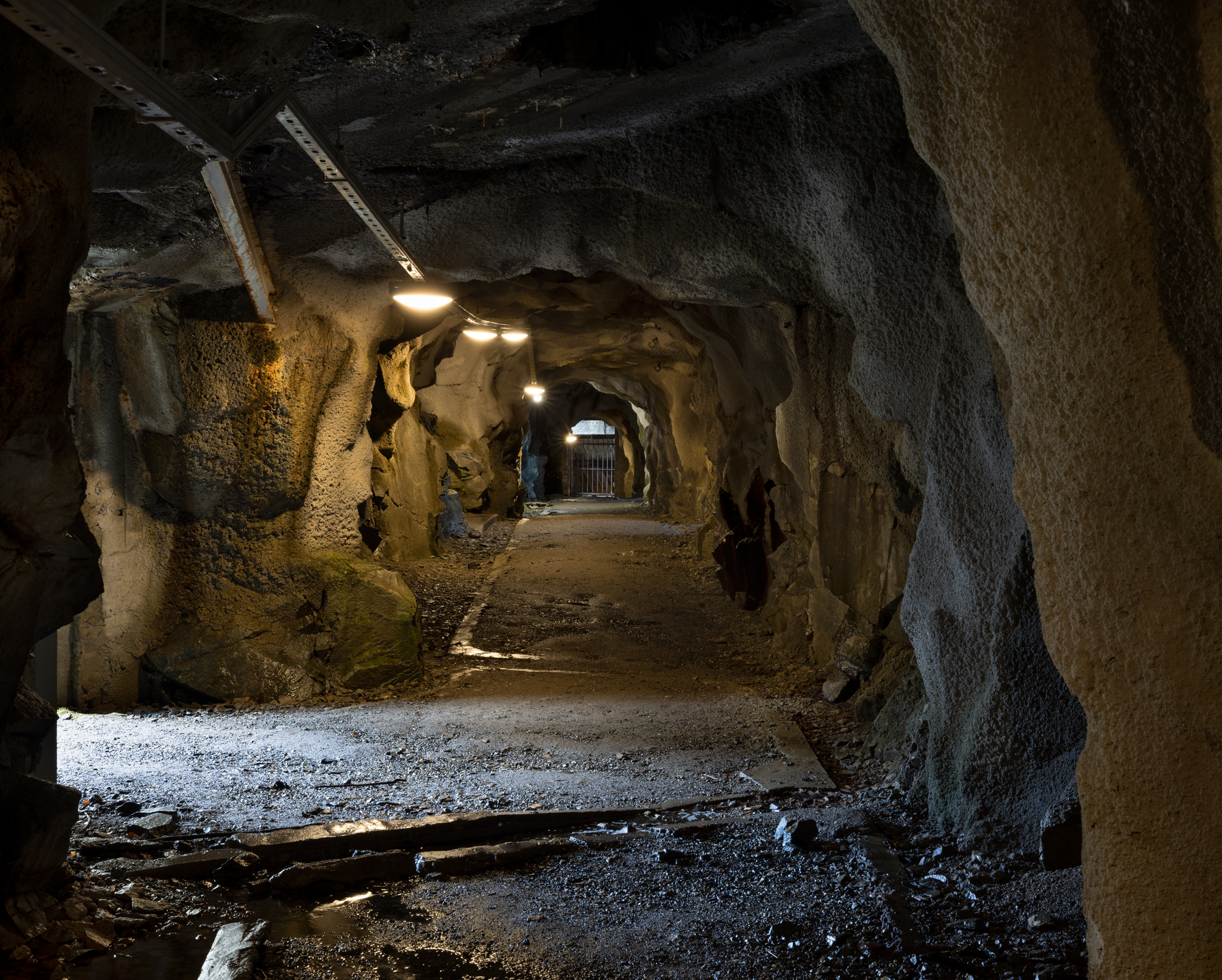 Svakt opplyst tunnel med grove steinvegger og -tak, opplyst av taklys, som fører til en metallport i den andre enden.
