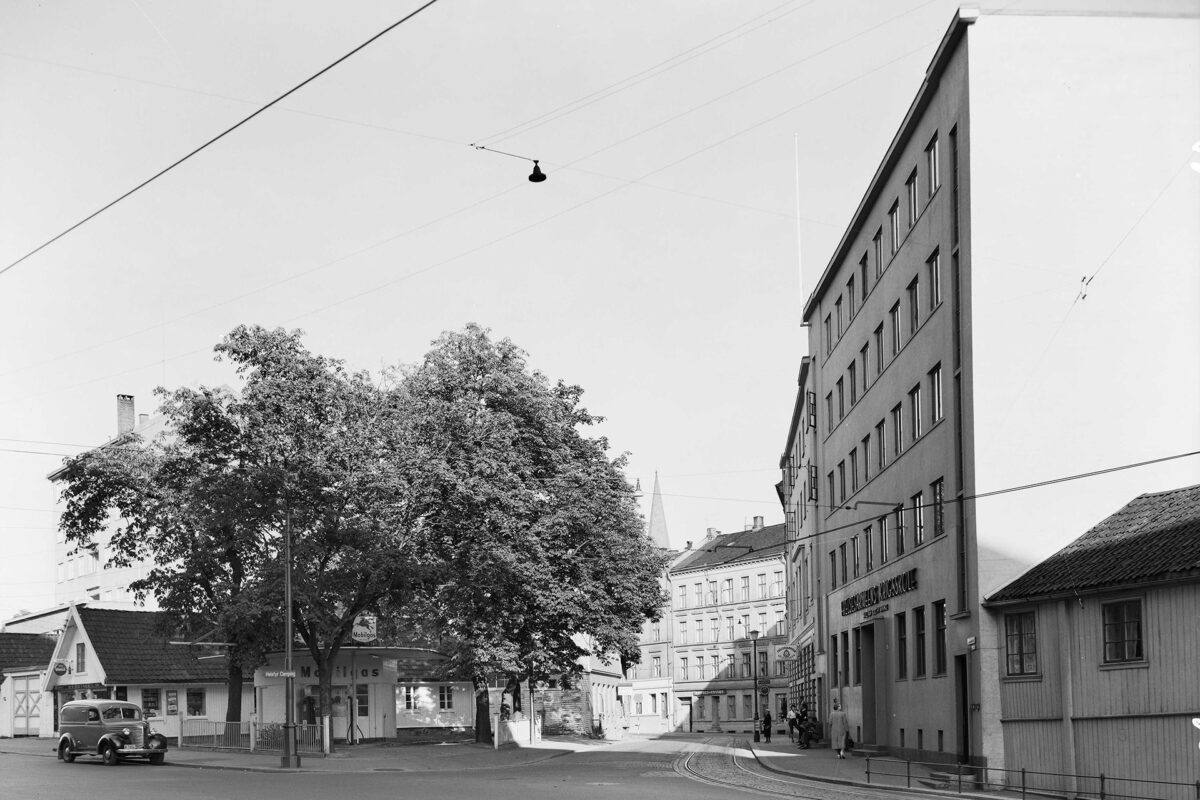 Svart-hvitt-bilde av et stille gatehjørne med flere trær, gamle biler og bygninger i flere etasjer, inkludert et skilt med teksten «Norsk Gjær- & Spritfabrikk».