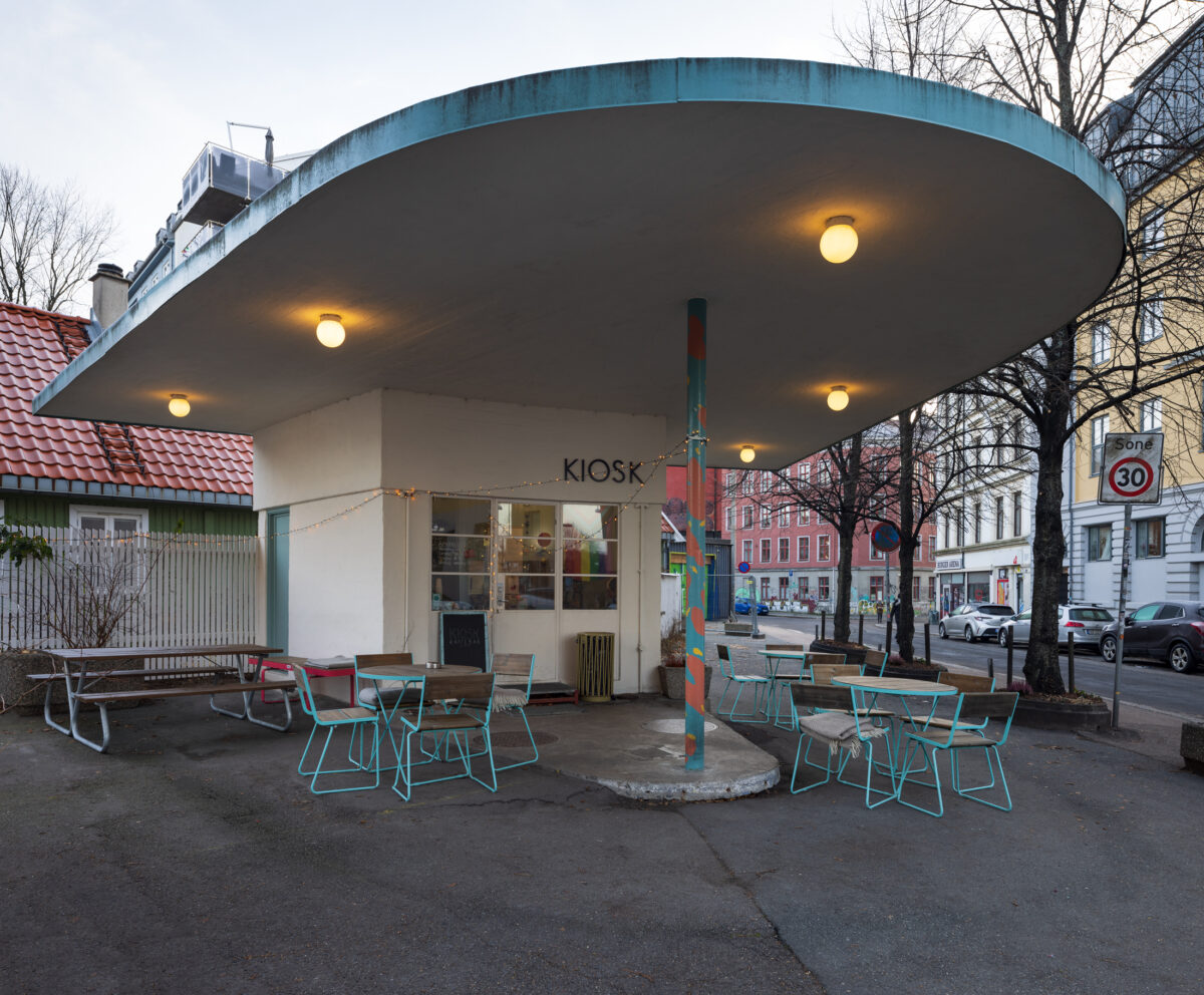 En liten kiosk med et stort overhengende tak, opplyst av runde lys, er omgitt av lyseblå utendørsbord og stoler på et tomt gatehjørne.