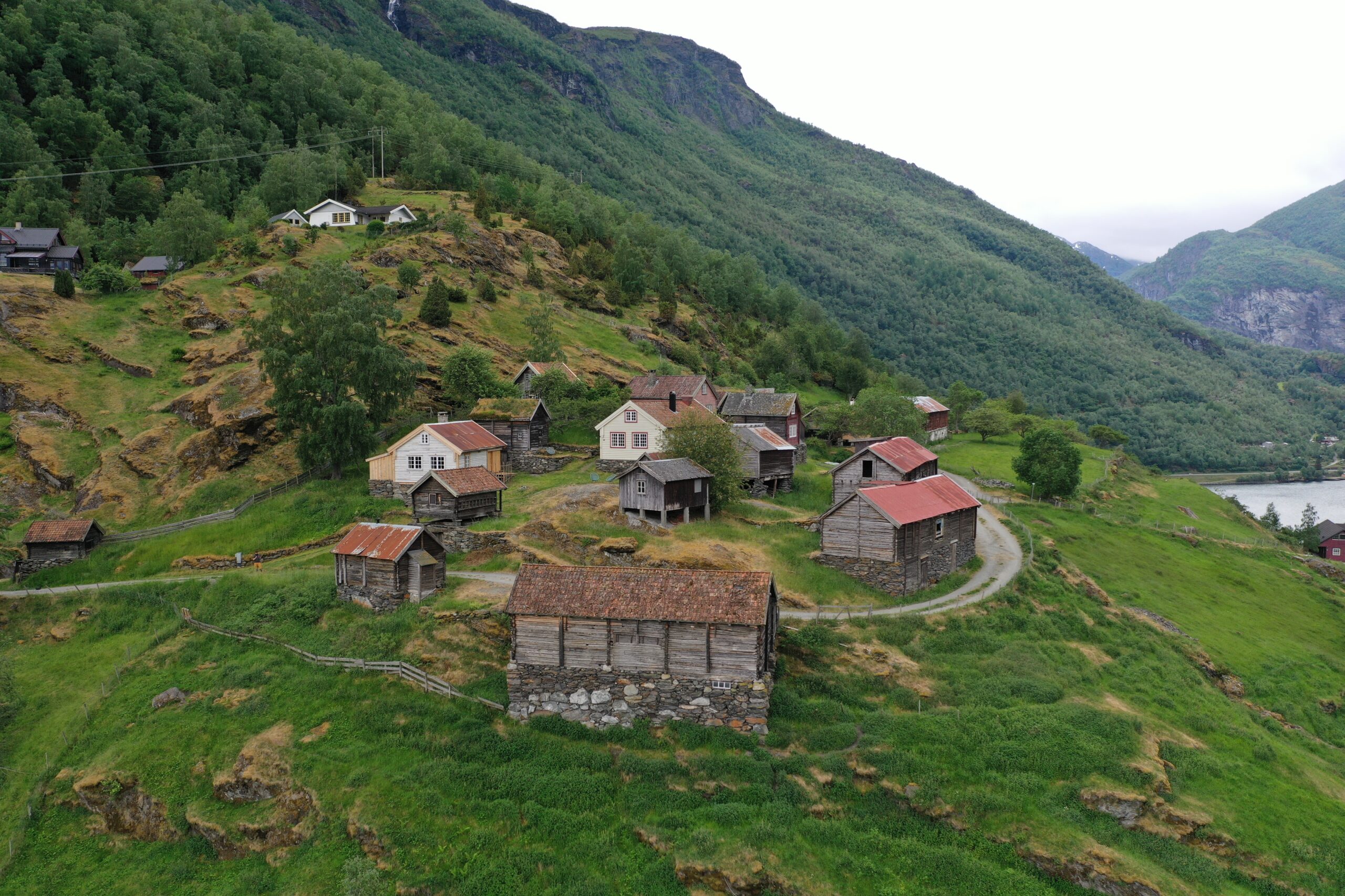En klynge av gamle tre- og steinbygninger på Otternes-tunet, ligger i en grønn åsside med en svingete sti, omgitt av frodige fjell og en elv i bakgrunnen.