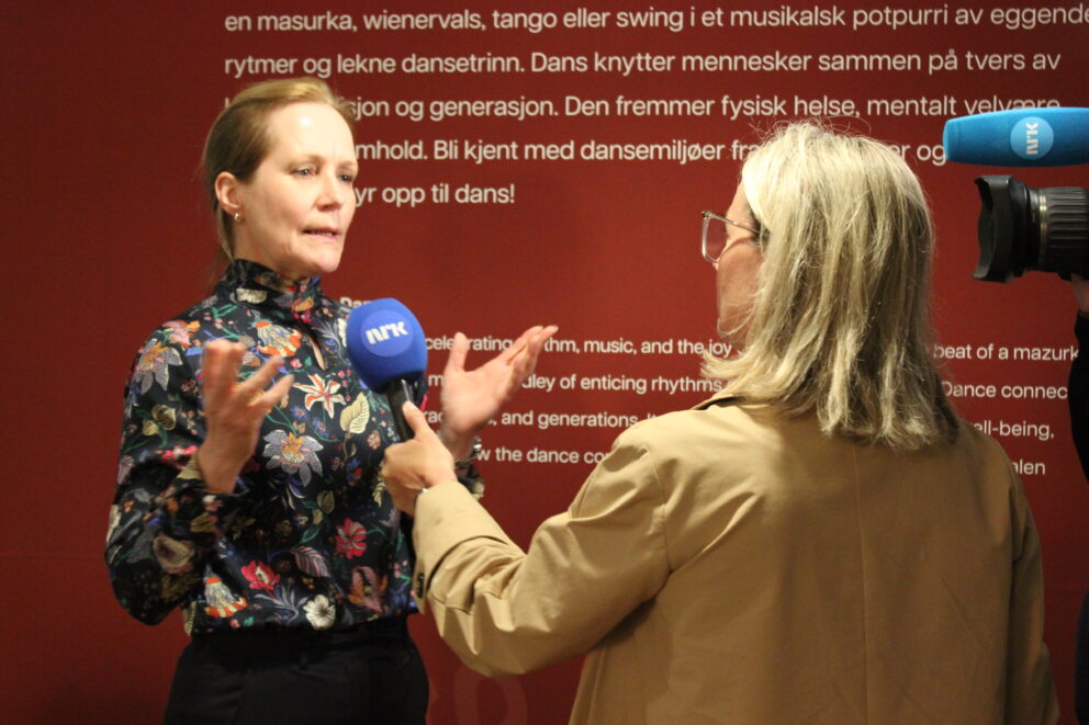 En kvinne blir intervjuet foran kamera av en journalist som holder en blå NRK-mikrofon foran en rød vegg med hvit tekst.