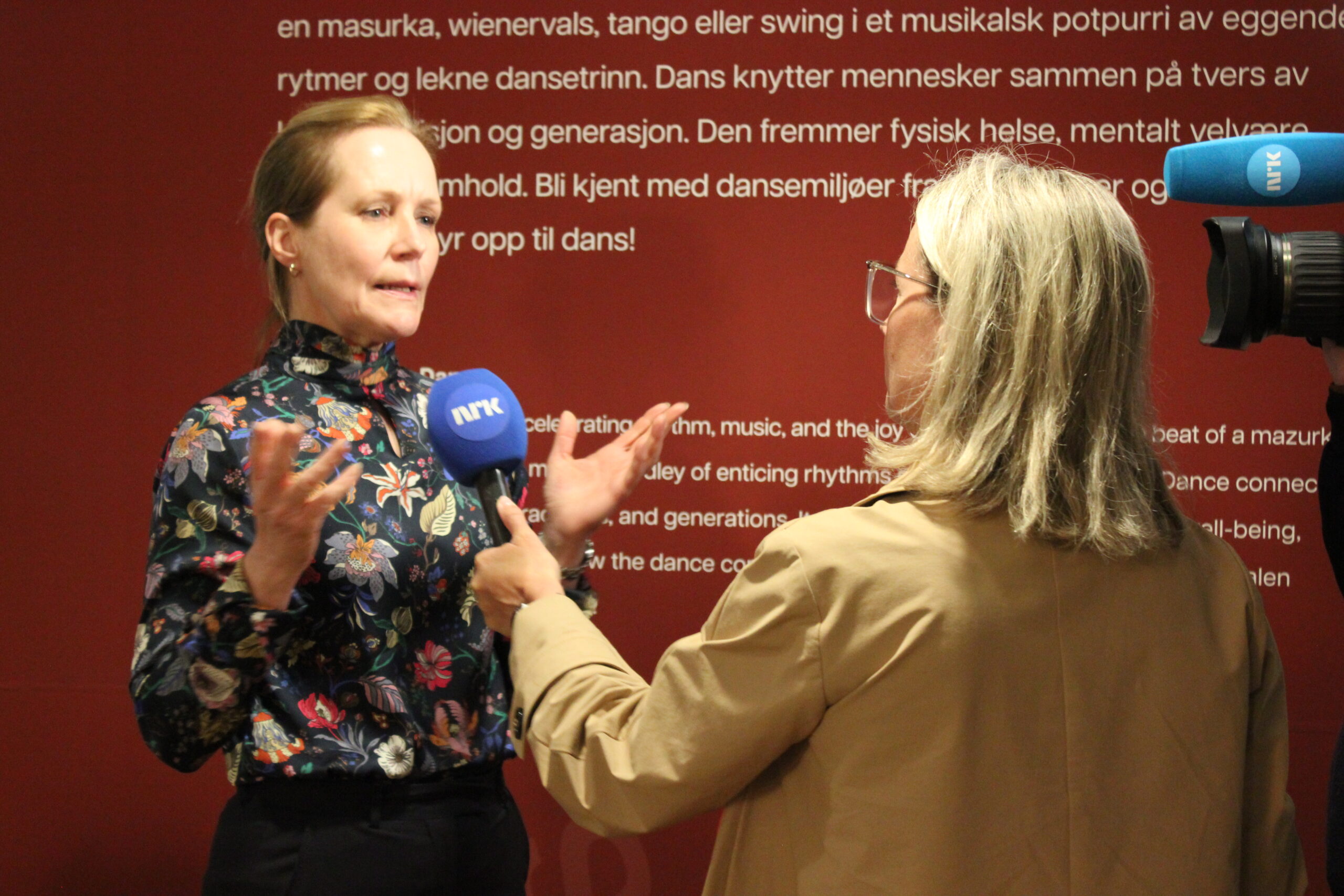 En kvinne blir intervjuet foran kamera av en journalist som holder en blå NRK-mikrofon foran en rød vegg med hvit tekst.