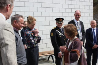 En gruppe mennesker, inkludert en uniformert offiser og formelt kledde personer, står utenfor nær en hvit murvegg og er i en samtale.