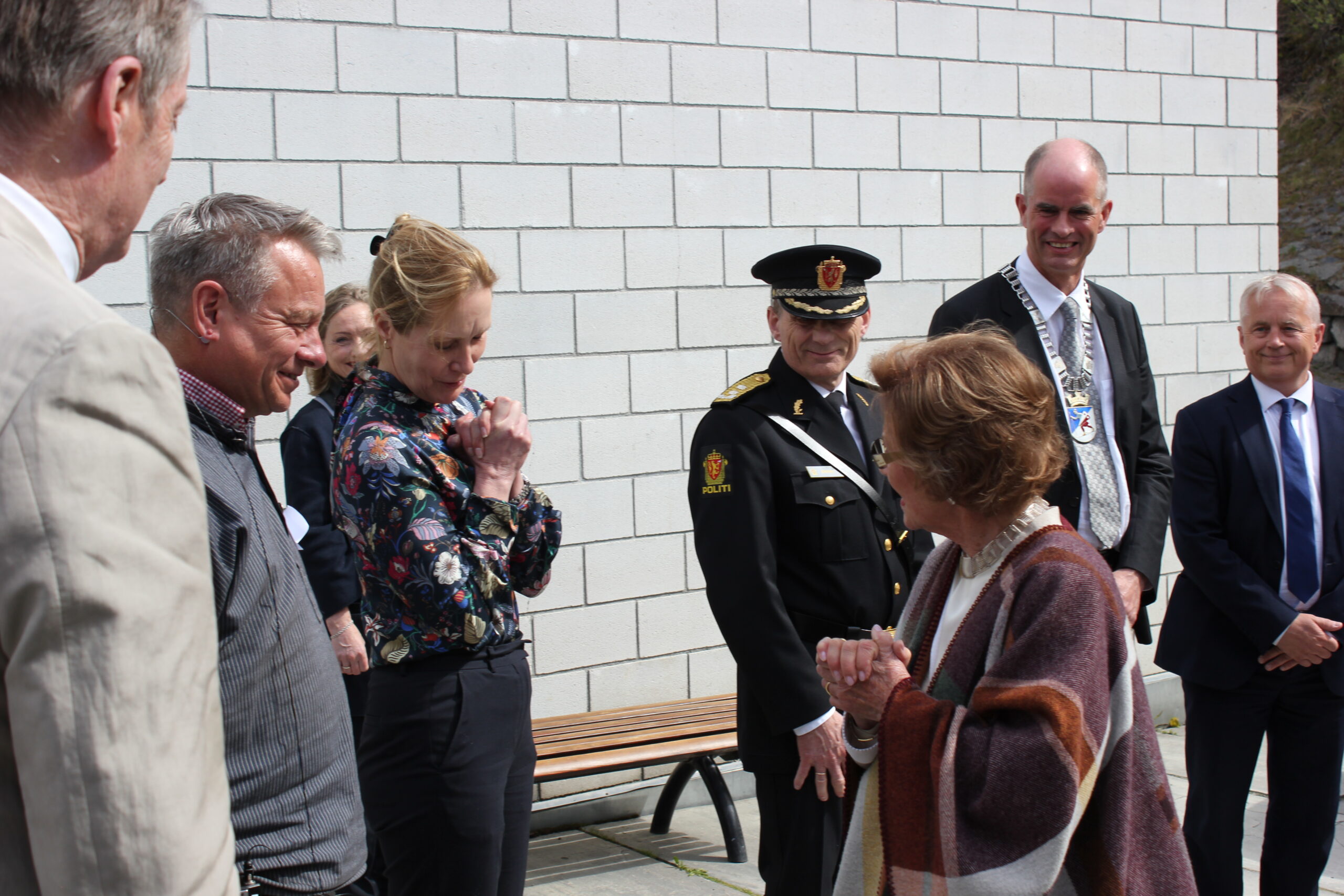 En gruppe mennesker, inkludert en uniformert offiser og formelt kledde personer, står utenfor nær en hvit murvegg og er i en samtale.