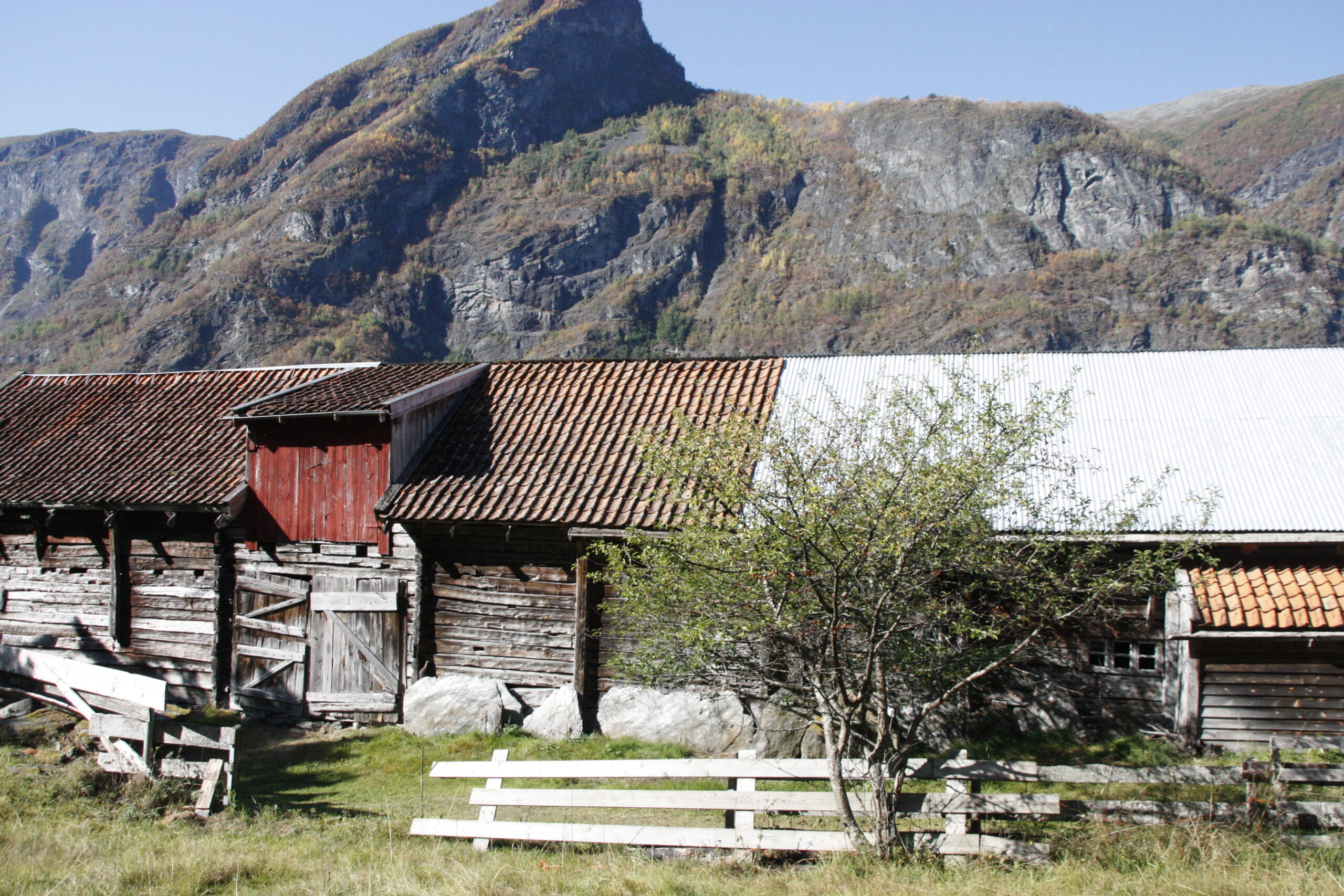 En gammel trelåve på Otternes-tunet med tegltak står foran et stort steinete fjell. Et tre og et ødelagt hvitt gjerde er i den gresskledde forgrunnen under en klar himmel.