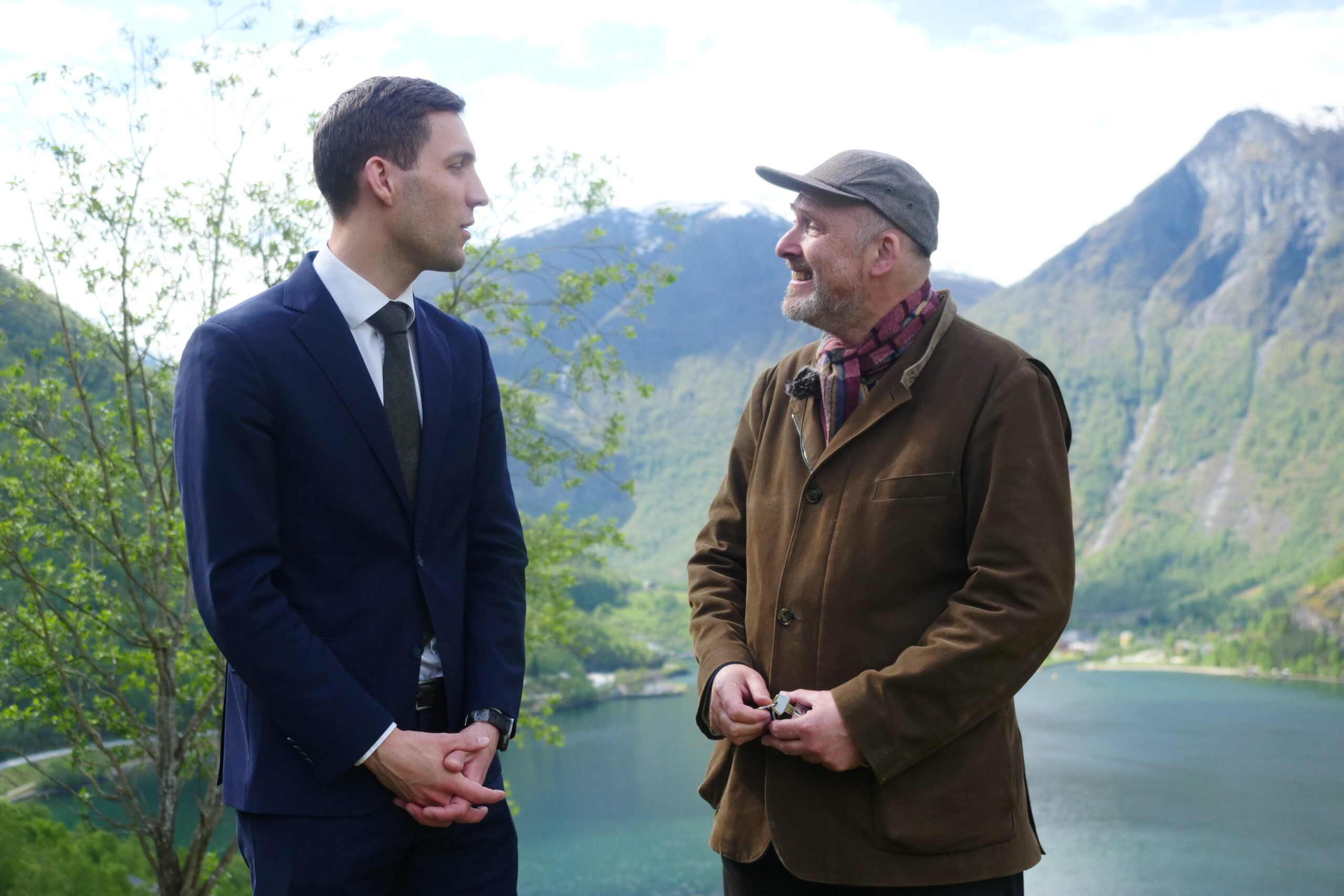 To menn står og snakker utendørs foran en innsjø og fjell; den ene har på seg dress og slips, den andre brun jakke og lue.