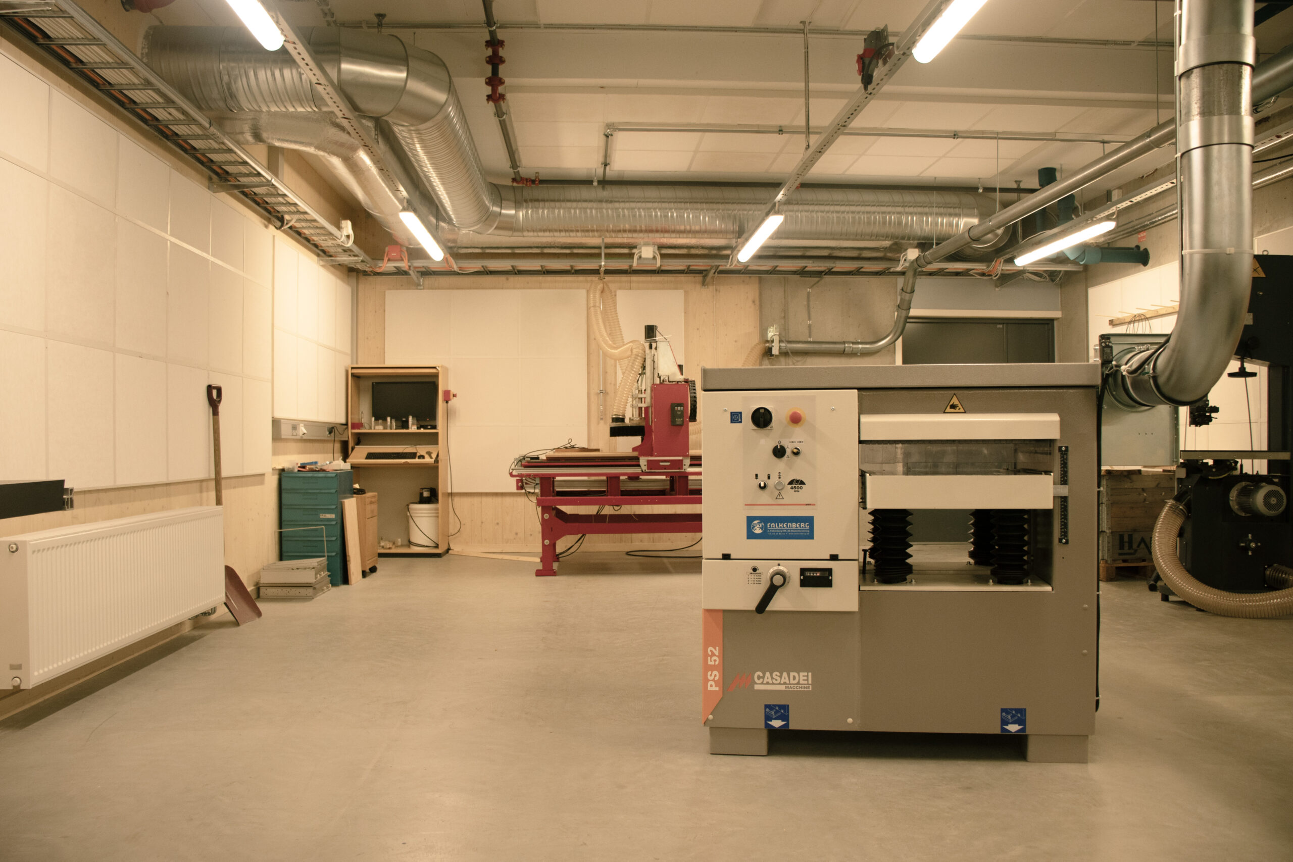 Et verksted med store industrielle trebearbeidingsmaskiner, ventilasjonskanaler i taket og verktøy organisert langs veggene.