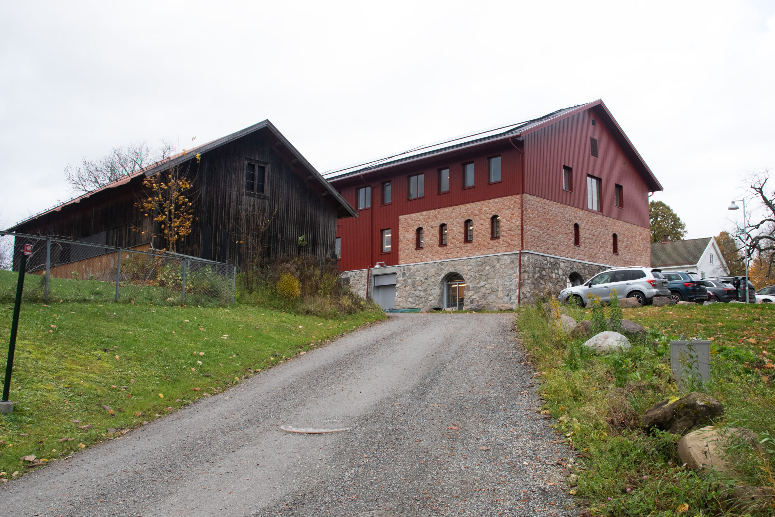 En gruslagt innkjørsel fører opp til en stor bygning i murstein og tre med flere parkerte biler på høyre side og en eldre trebygning på venstre side.