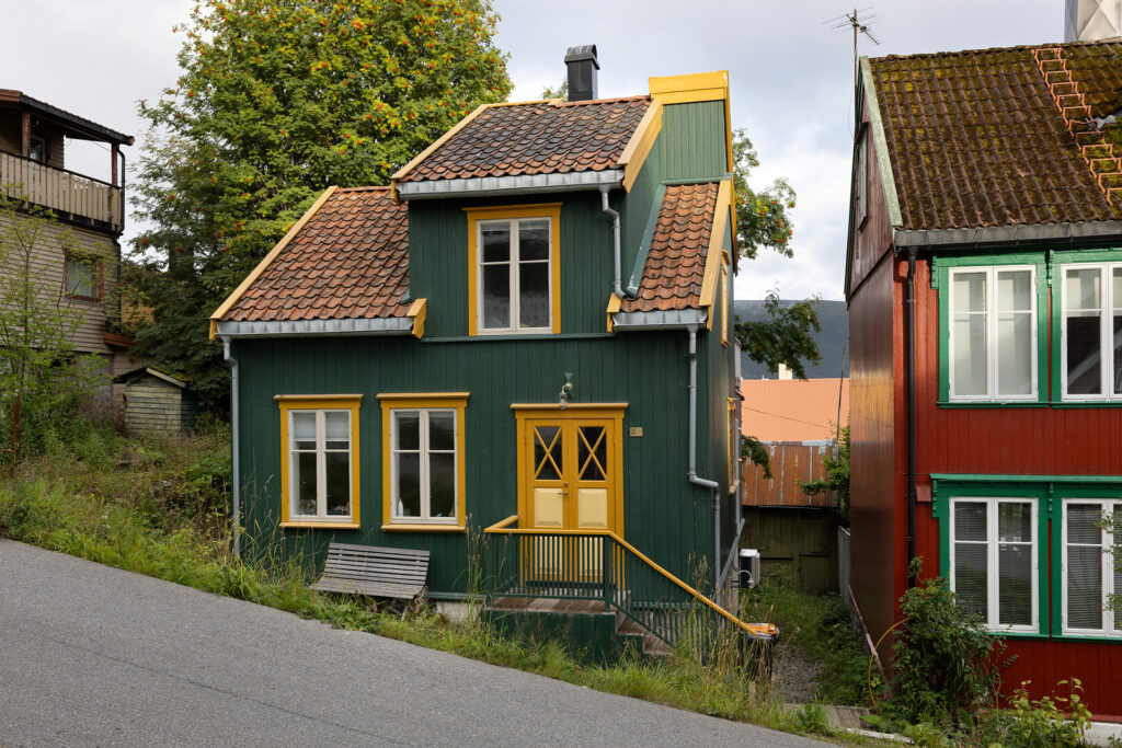 Et lite, grønt trehus med gule kanter og rødt tegltak ligger i en skrå gate, ved siden av et rødt hus med hvite vinduer.