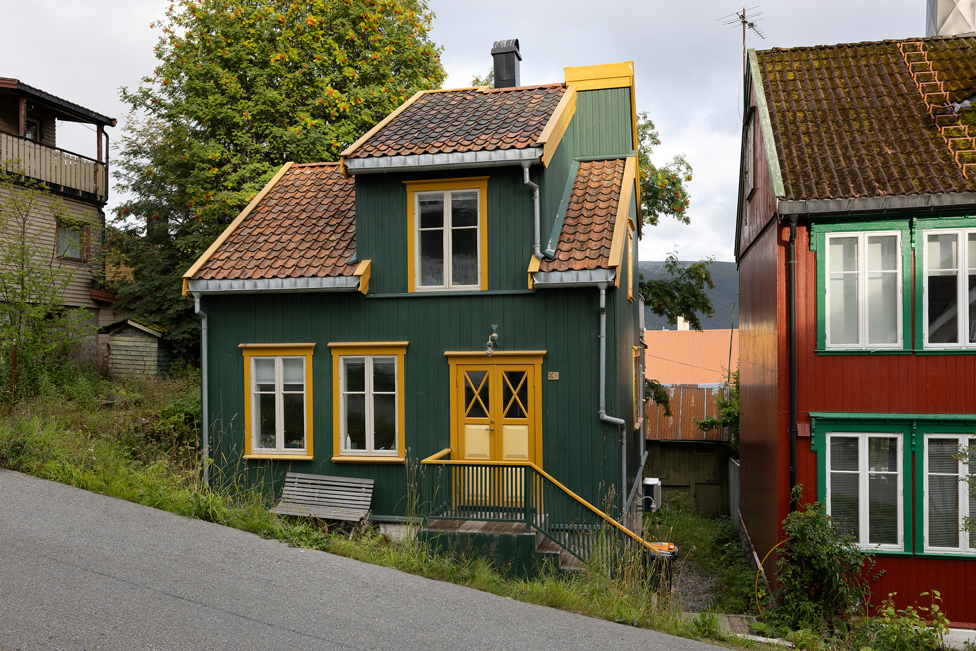 Et lite, grønt trehus med gule kanter og rødt tegltak ligger i en skrå gate, ved siden av et rødt hus med hvite vinduer.
