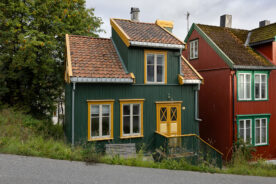 Et lite, grønt trehus med gule kanter og tegltak står ved siden av et rødt hus i en skrånende gate med gress og trær i nærheten.