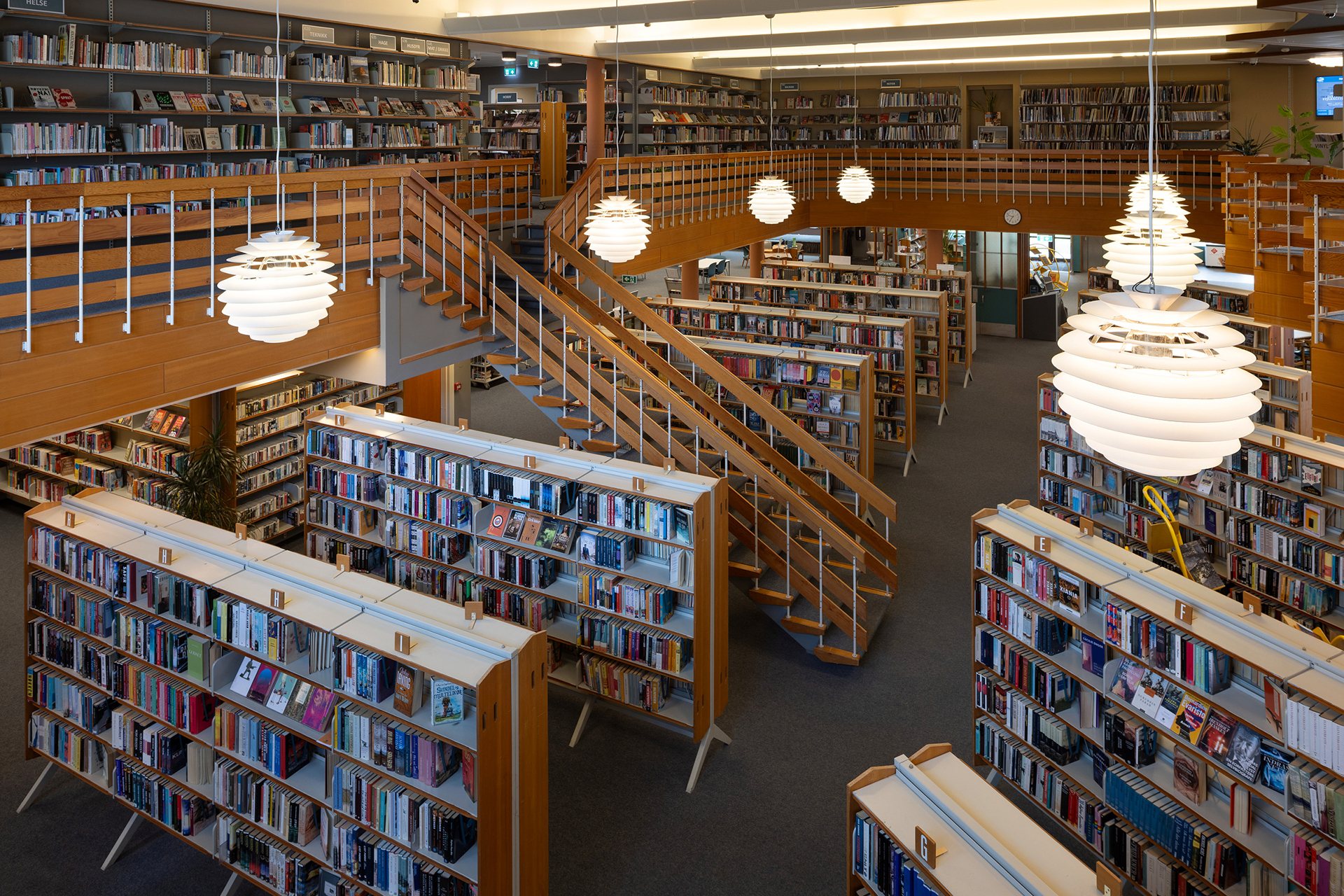 Et romslig bibliotekinteriør med bokhyller i tre, flere rader med bøker, en sentral trapp og moderne runde pendellamper som henger fra taket.