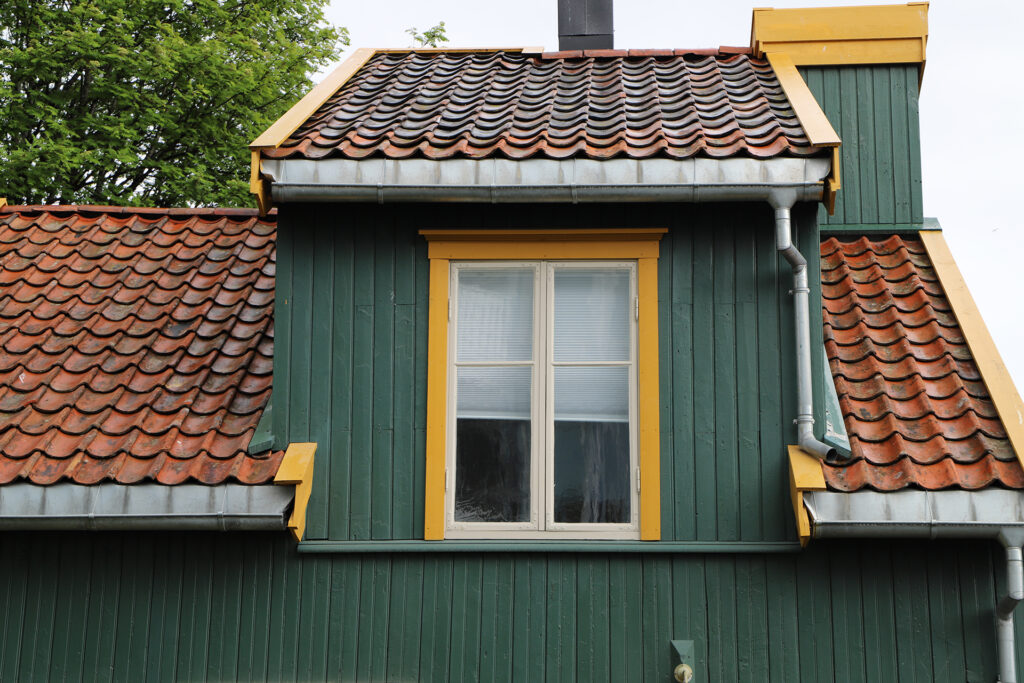 Et grønt trehus med rødt tegltak, et sentralt vindu med gul kant og synlige takrenner langs kantene.