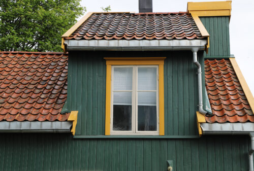 Et grønt trehus med rødt tegltak, et sentralt vindu med gul kant og synlige takrenner langs kantene.