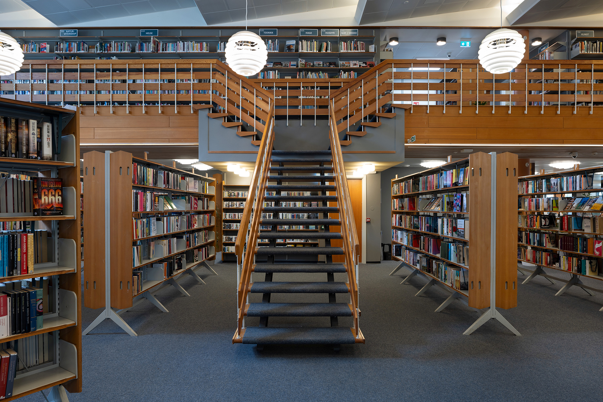 En trapp fører opp til en mesanin i et moderne bibliotek, omgitt av bokhyller fylt med bøker.