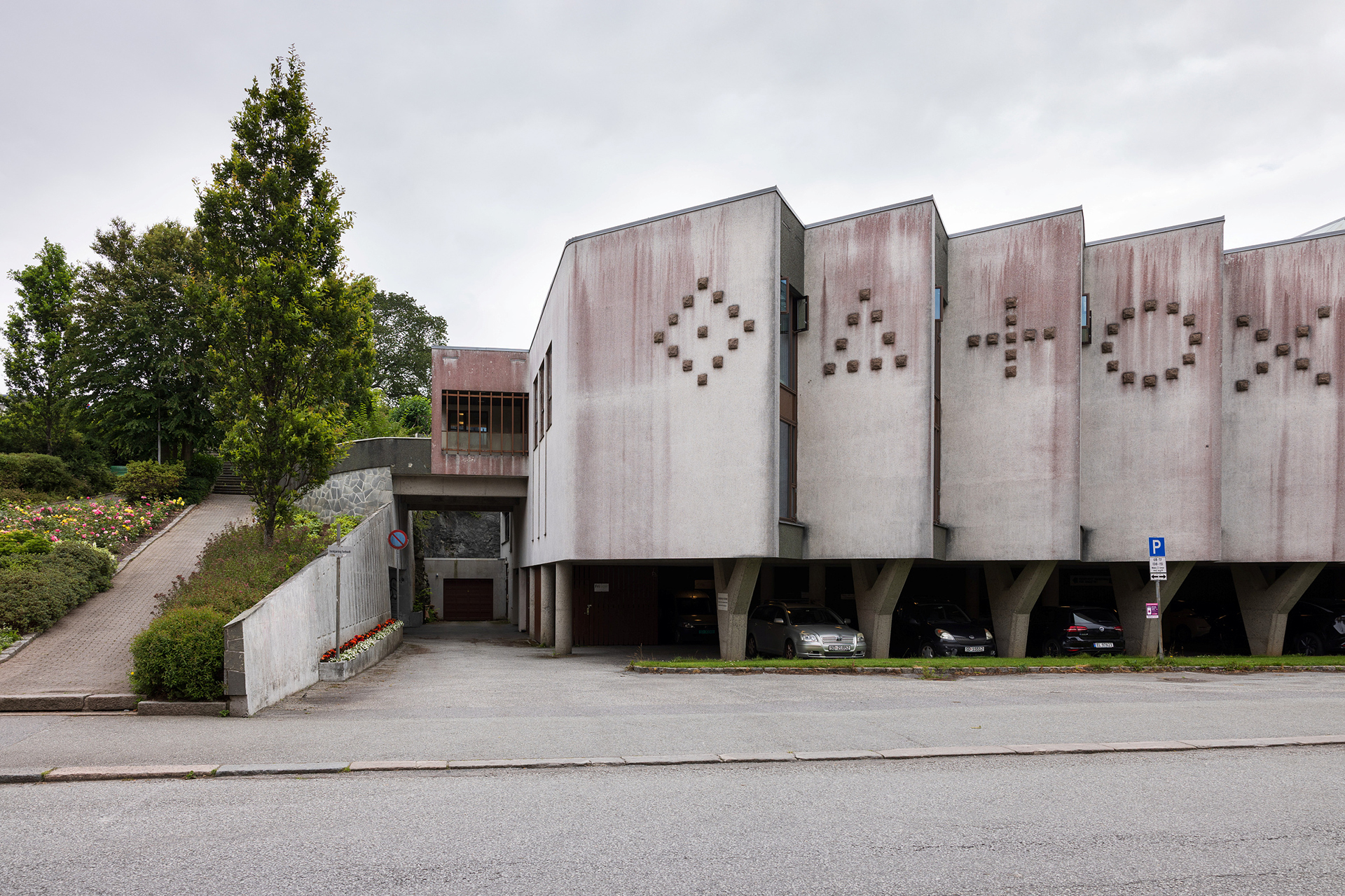 Bygning i brutalistisk stil med geometriske former, støttet av søyler, og biler parkert under; et mønster av prikker danner symboler på fasaden.