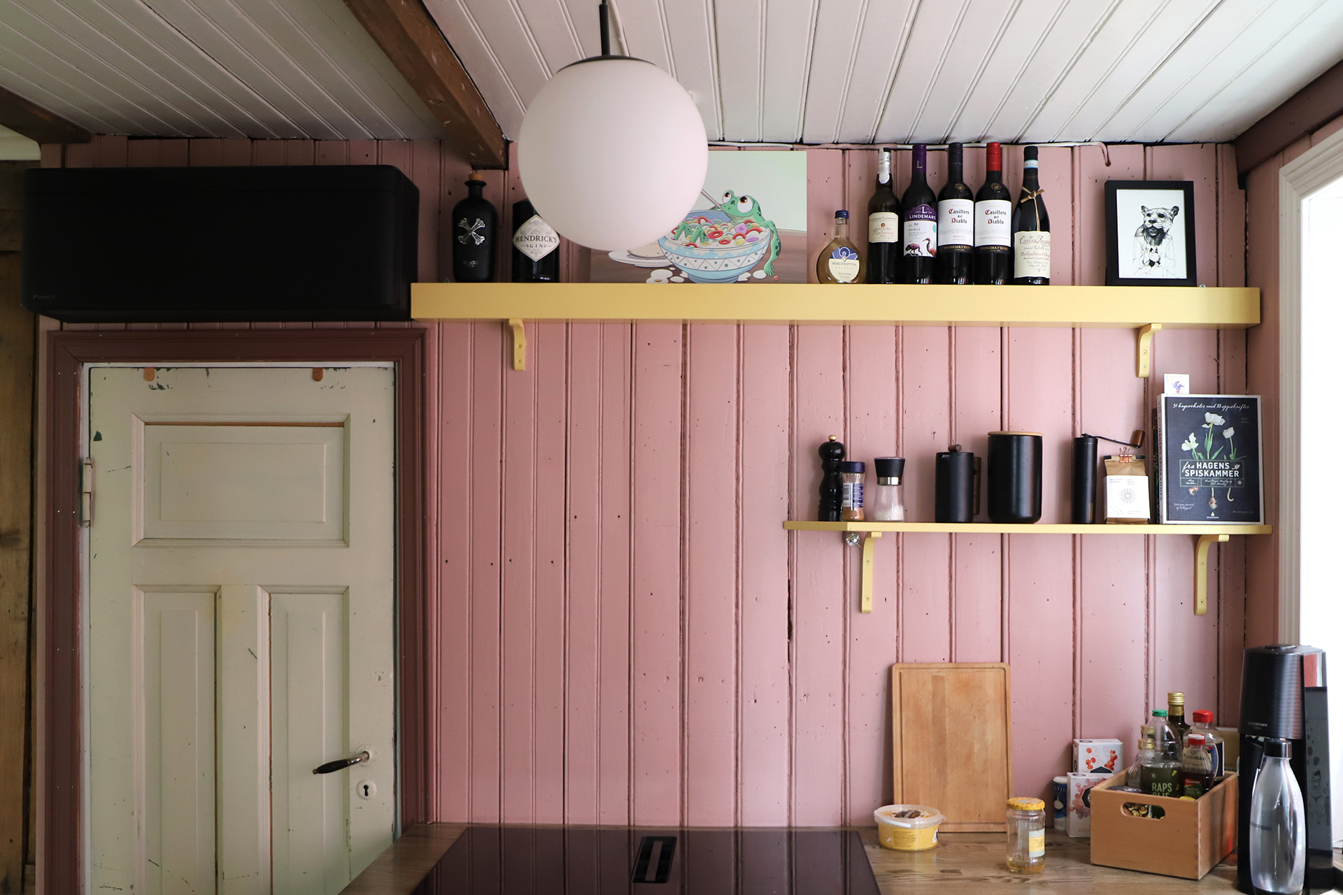 Et lite kjøkken med rosa trevegger, to gule hyller med flasker, kopper og dekor, en lukket dør og diverse kjøkkenting på benken.