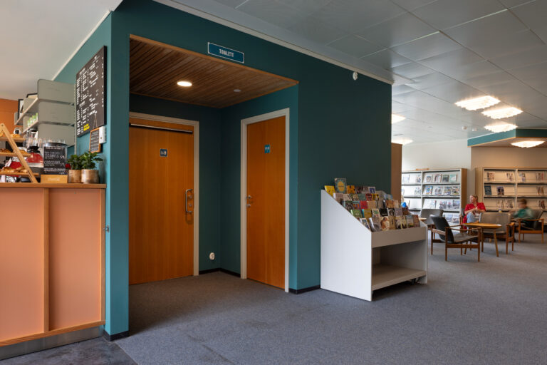 Interiør i et bibliotek eller offentlig rom med toalettdører, et brosjyrestativ, stoler, bord og bokhyller i bakgrunnen.