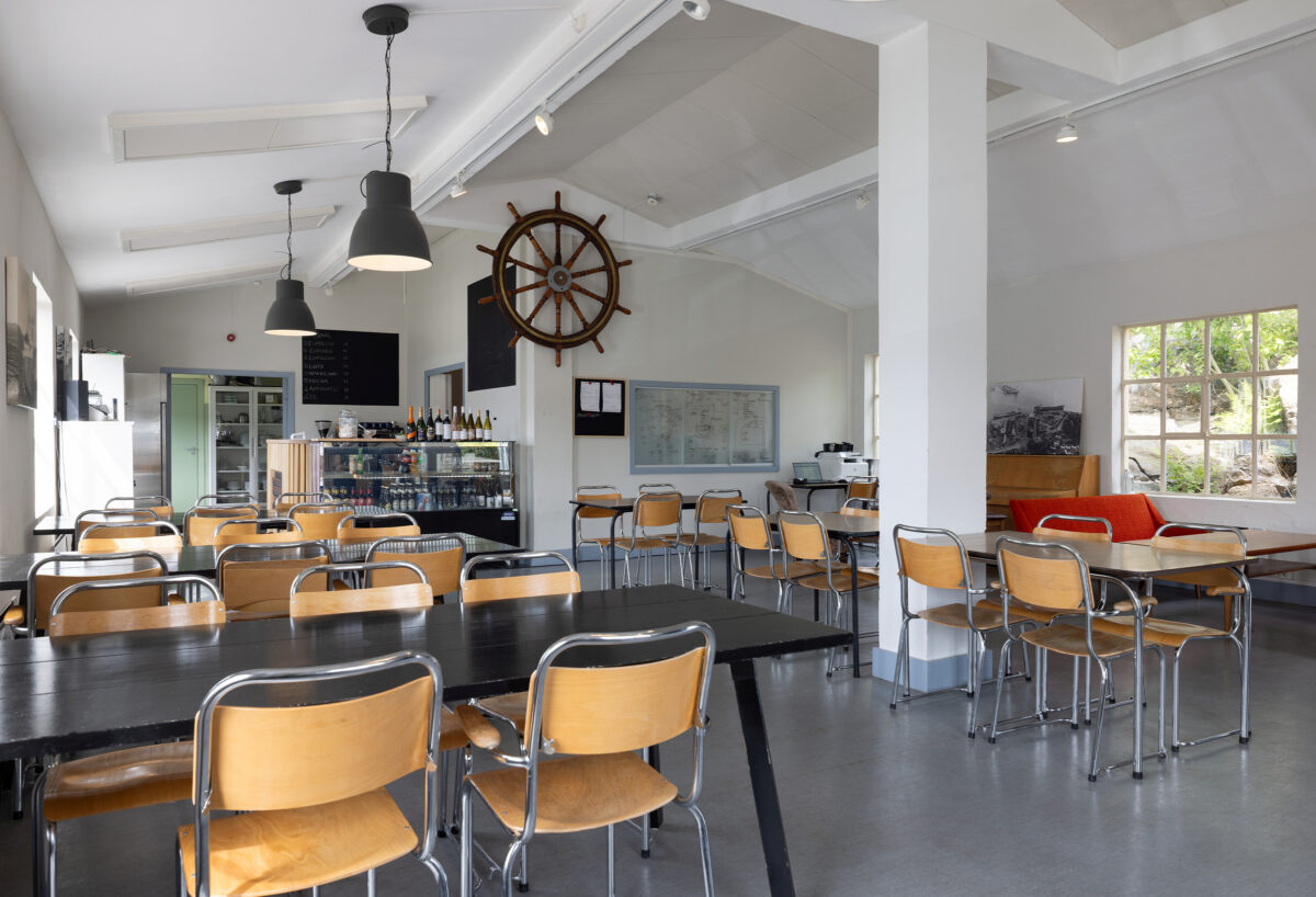 Et lyst og moderne kaféinteriør med trestoler, svarte bord, en disk med mat og drikke, nautisk innredning og store vinduer som slipper inn naturlig lys.