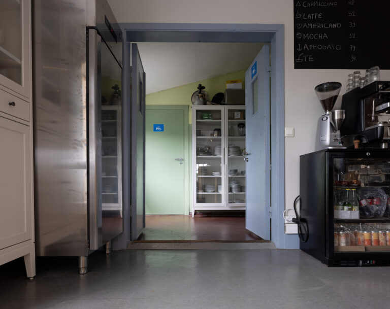 Utsikt over et kjøkken med blå dører, hyller med servise, kjøleskap, kaffemaskin og en menytavle på veggen.
