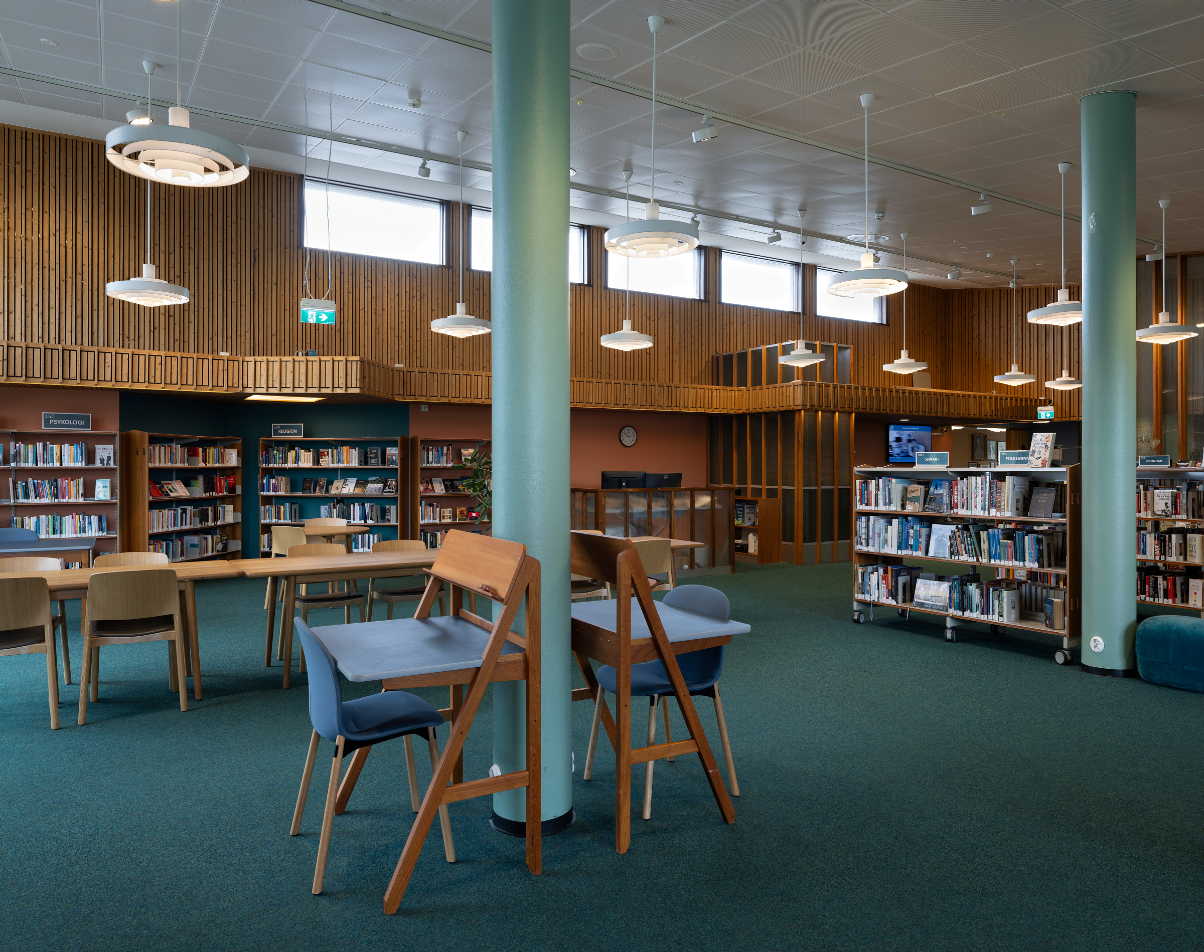 Moderne bibliotekinnredning med bokhyller, bord, stoler, grønt teppe, trepanel og taklamper. Rommet er tomt og godt opplyst med naturlig og kunstig lys.