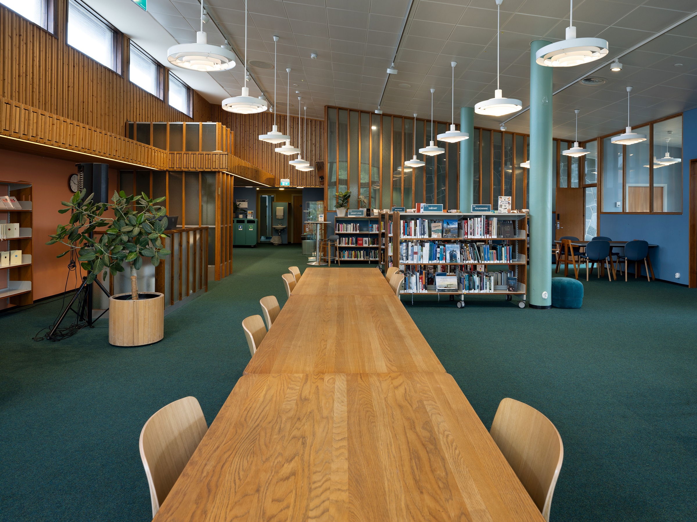 Et romslig bibliotek med bord og stoler i tre, bokhyller, pendellamper, grønne tepper og store vinduer som slipper inn naturlig lys.