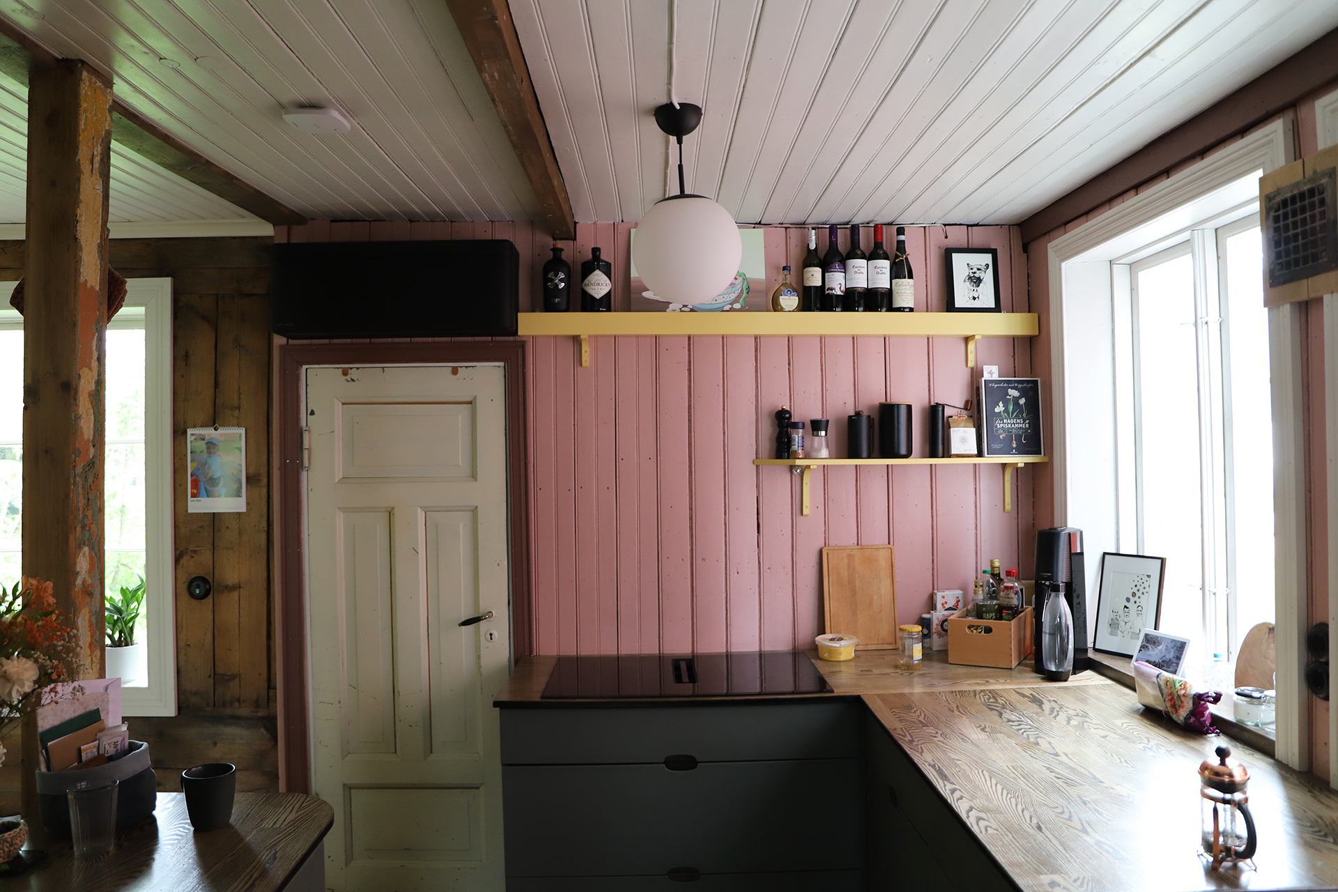 Et kjøkken med rosa trevegger, en benk i tre, svarte skap, åpne hyller med flasker og dekor, og to vinduer som slipper inn naturlig lys.