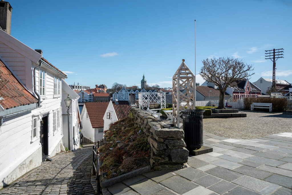 En brosteinsbelagt gate og hvite trehus med røde tak, en steinmur, benker og et løvløst tre under en klarblå himmel i en stille by.