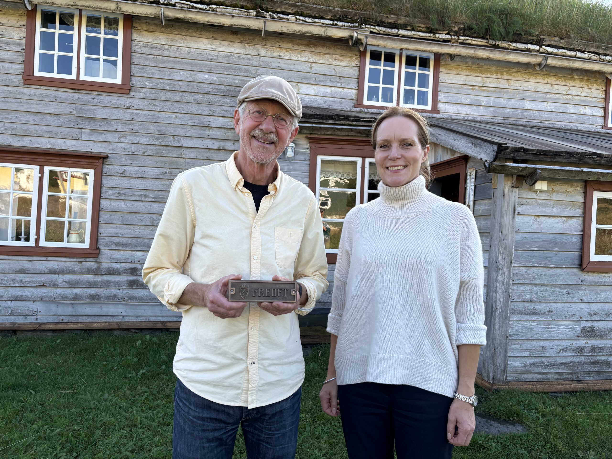 To personer står smilende foran en trebygning med gresstak; personen til venstre holder en liten plakett eller murstein.
