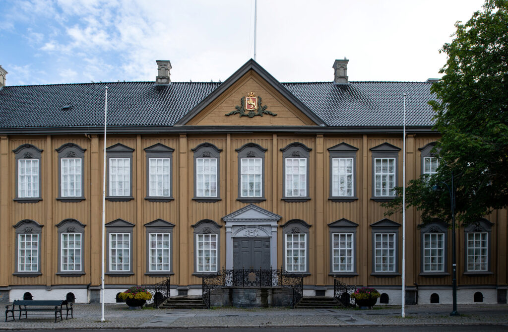 En symmetrisk trebygning i to etasjer med mange hvitkalkede vinduer, en sentral inngang og et dekorativt våpenskjold over døren. To tomme flaggstenger står foran.