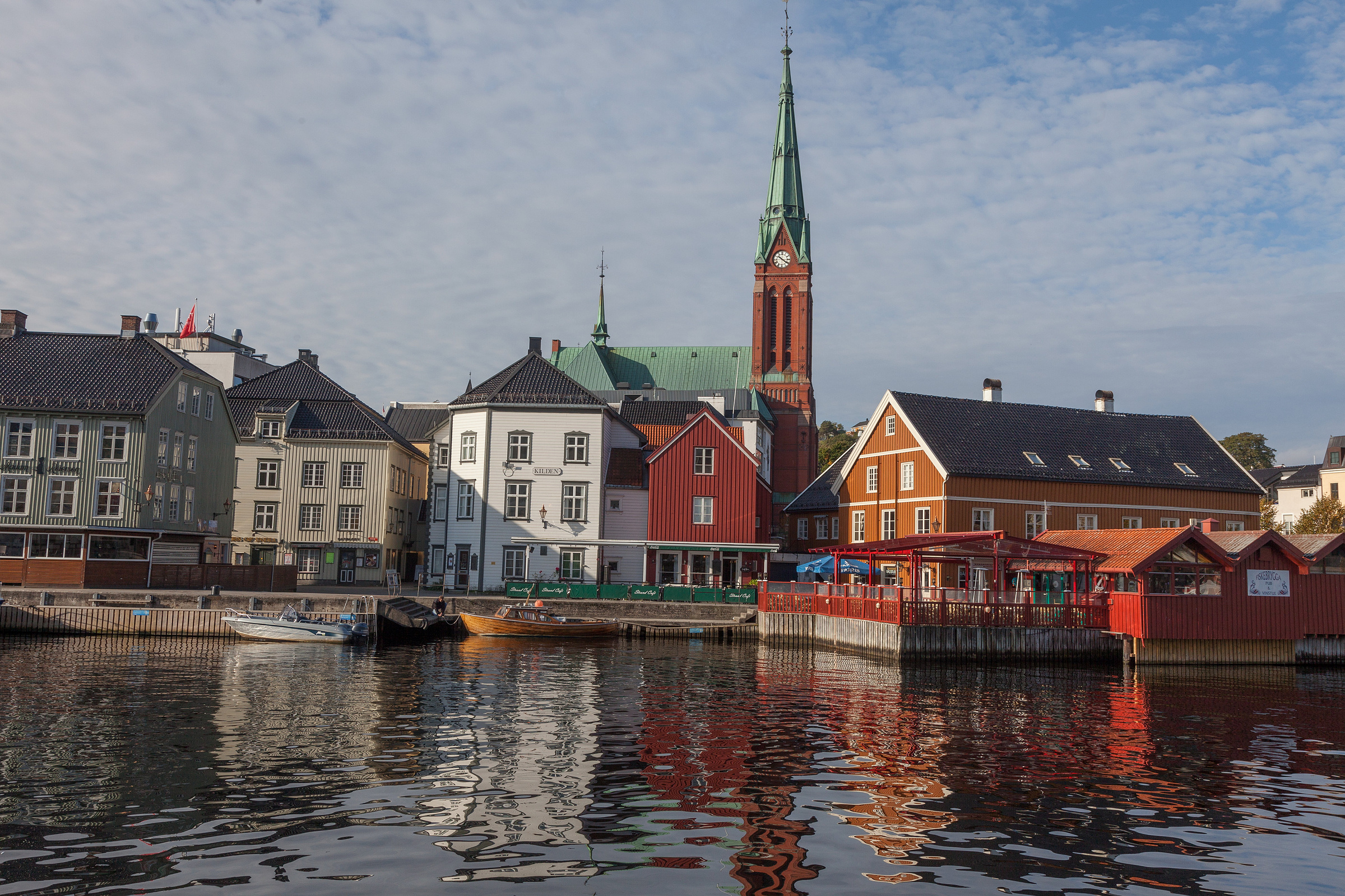 En rekke fargerike bygninger og båter ved vannkanten med en stor mursteinskirke og et høyt spir i bakgrunnen, som speiler seg i det rolige vannet under en delvis overskyet himmel.