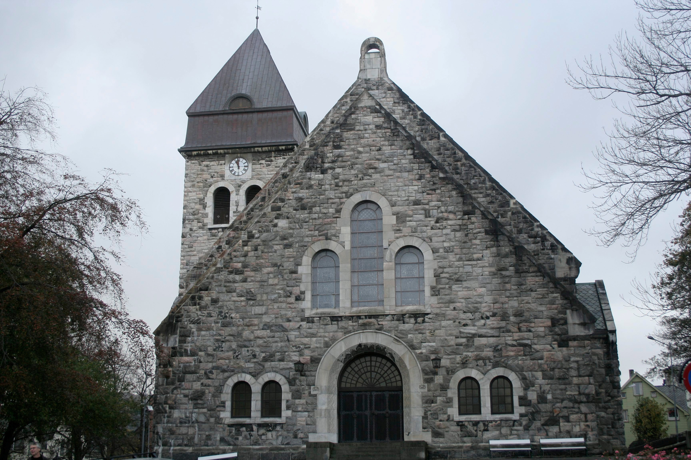 Ålesund er ei steinkirke med buede vinduer og dører, klokketårn med klokke og trær på hver side, fotografert en overskyet dag. Den har fått kyrkjemidlar.